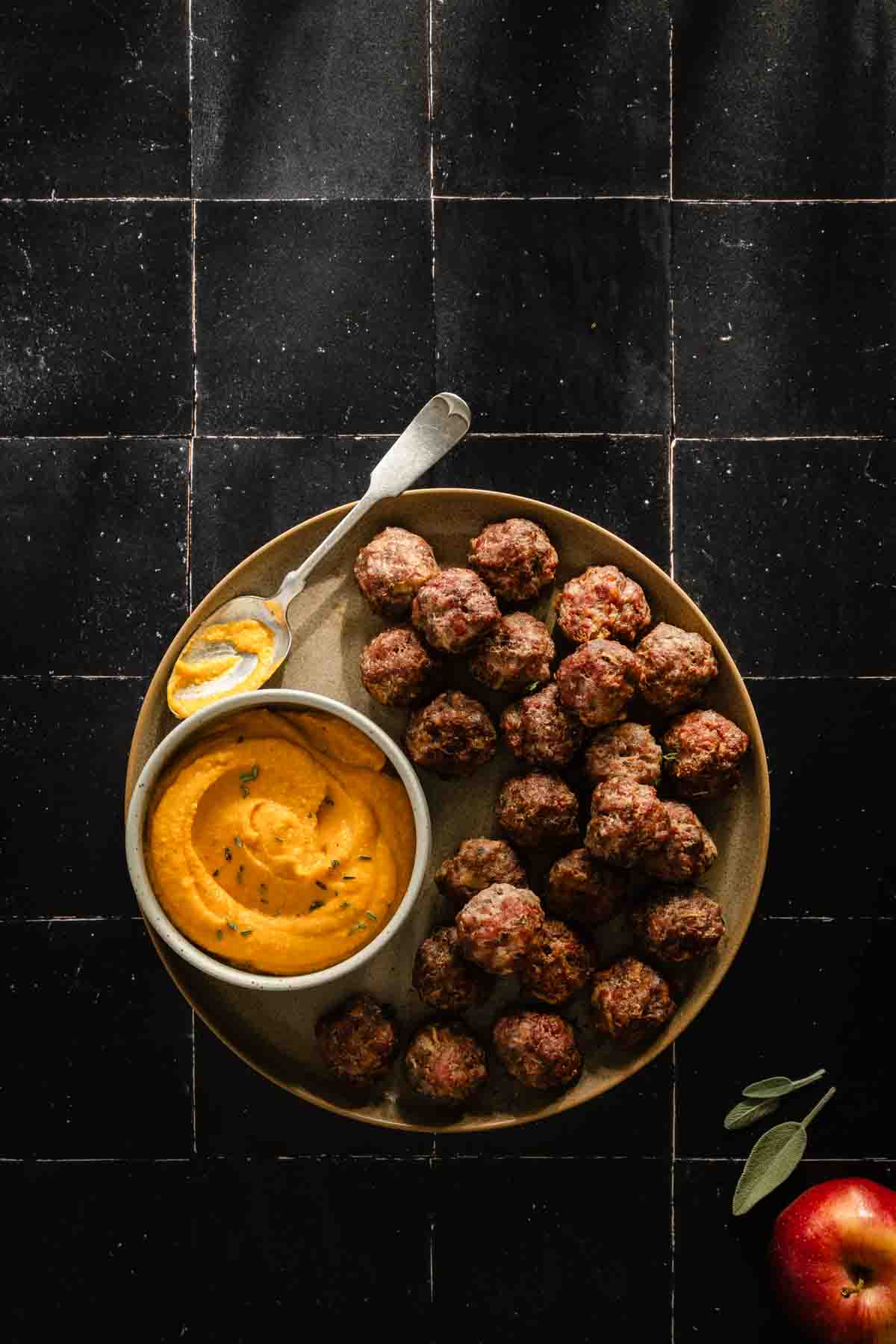 meatballs on a round plate with a side of sauce; dark background; apple and few sage leaves in the lower right corner