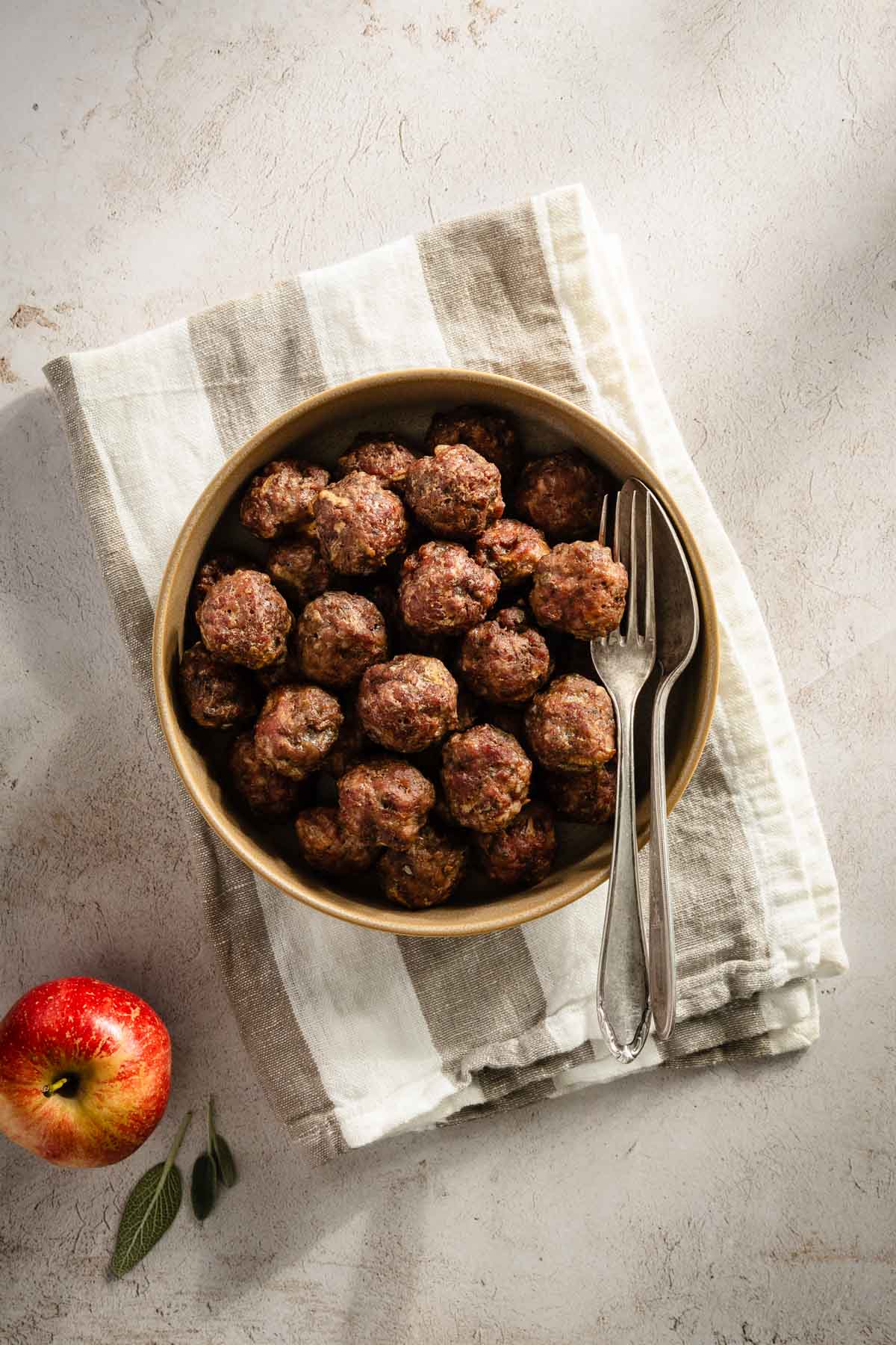 cooked meatballs in a glass bowl, apple and few sage leaves in the lower left corner