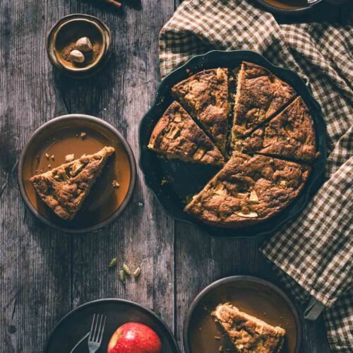 Slice of brown butter apple cake on a brown plate with a fork, black coffee mug, teal linen, and cinnamon sticks on rustic wood.