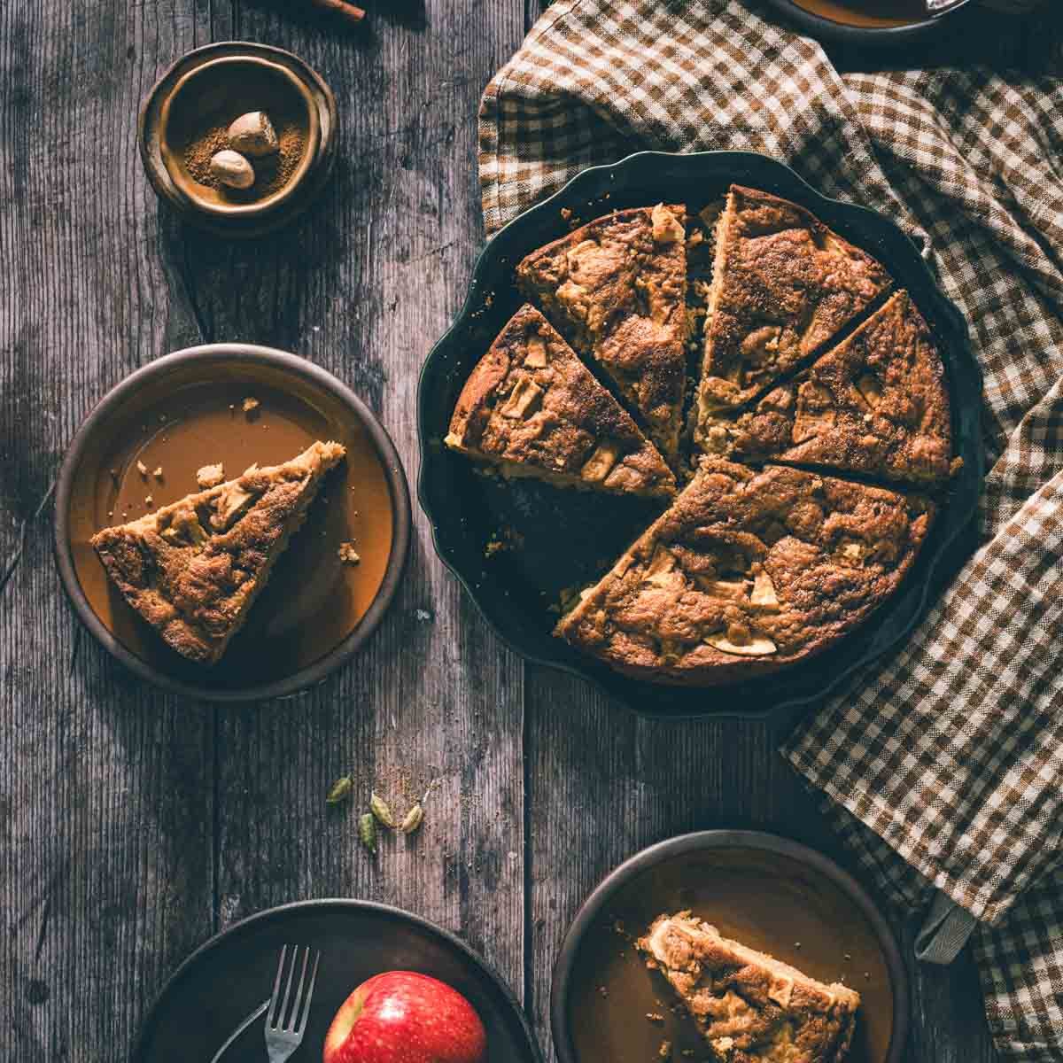 Slice of brown butter apple cake on a brown plate with a fork, black coffee mug, teal linen, and cinnamon sticks on rustic wood.