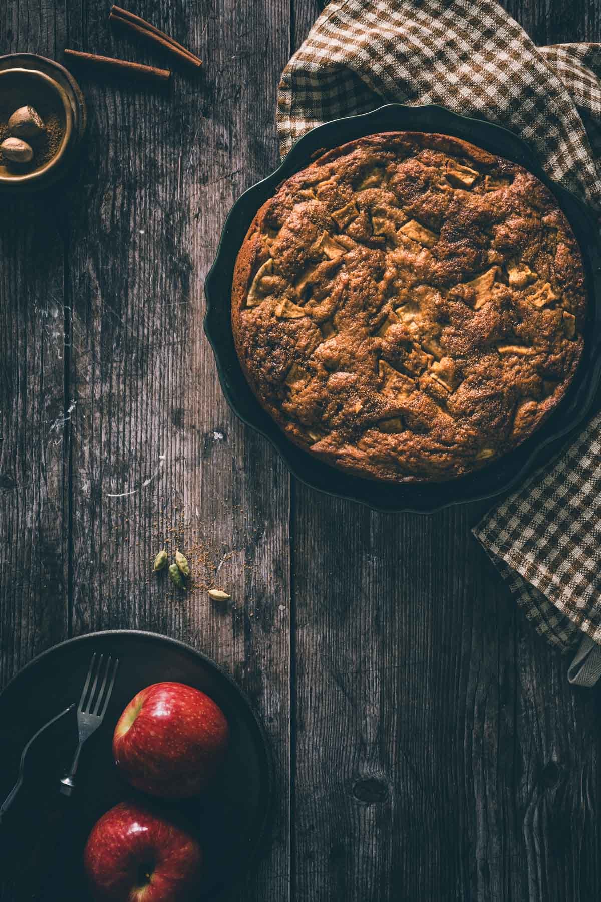 Top view of a whole caramelized apple cake in a green dish with a brown gingham towel, forks, and cinnamon sticks.