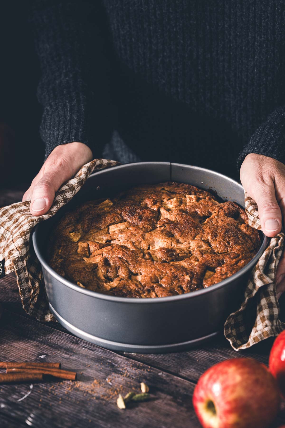 Hands holding a freshly baked brown butter apple cake in a springform pan with a gingham towel, red apples nearby.