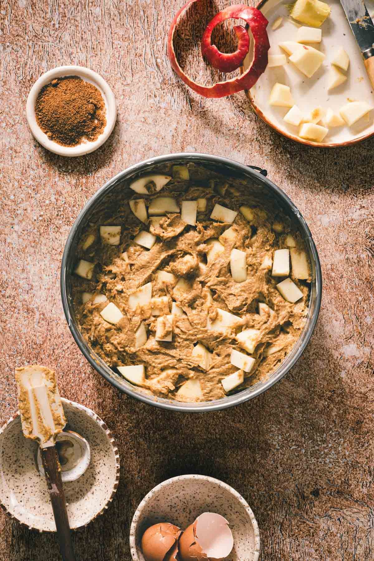 Apple cake batter in a springform pan surrounded by diced apples, apple peel, spiced sugar, a spatula, and eggshells.