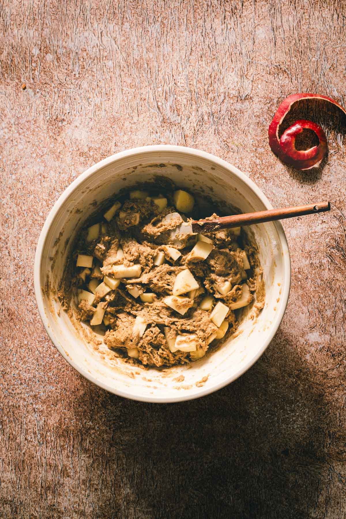 One-bowl brown butter cake batter with folded-in apple chunks, a wooden spatula, and a curl of red apple peel.