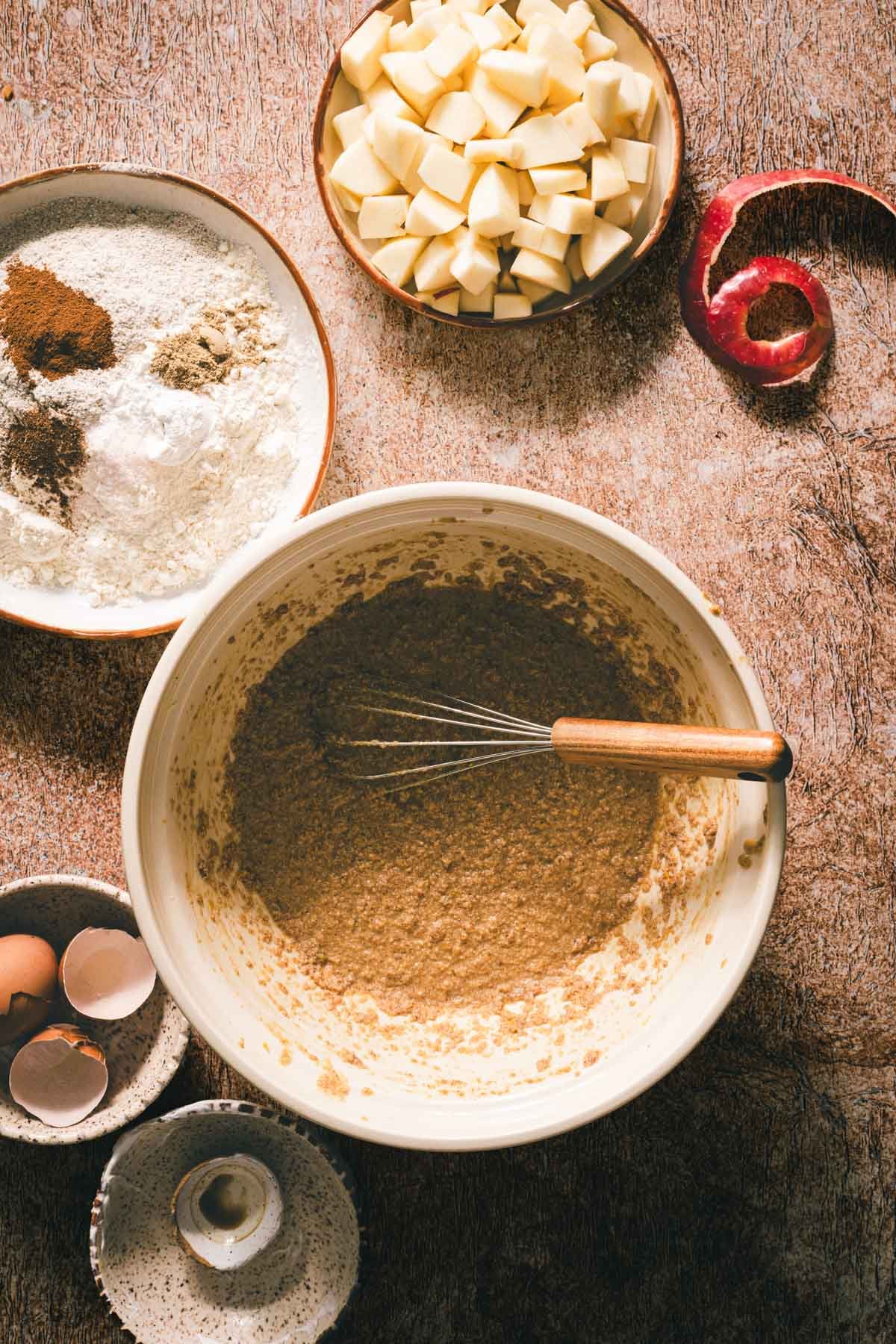 Whisking brown butter, sugar, and eggs in one bowl, beside diced apples and flour mixed with warm spices.