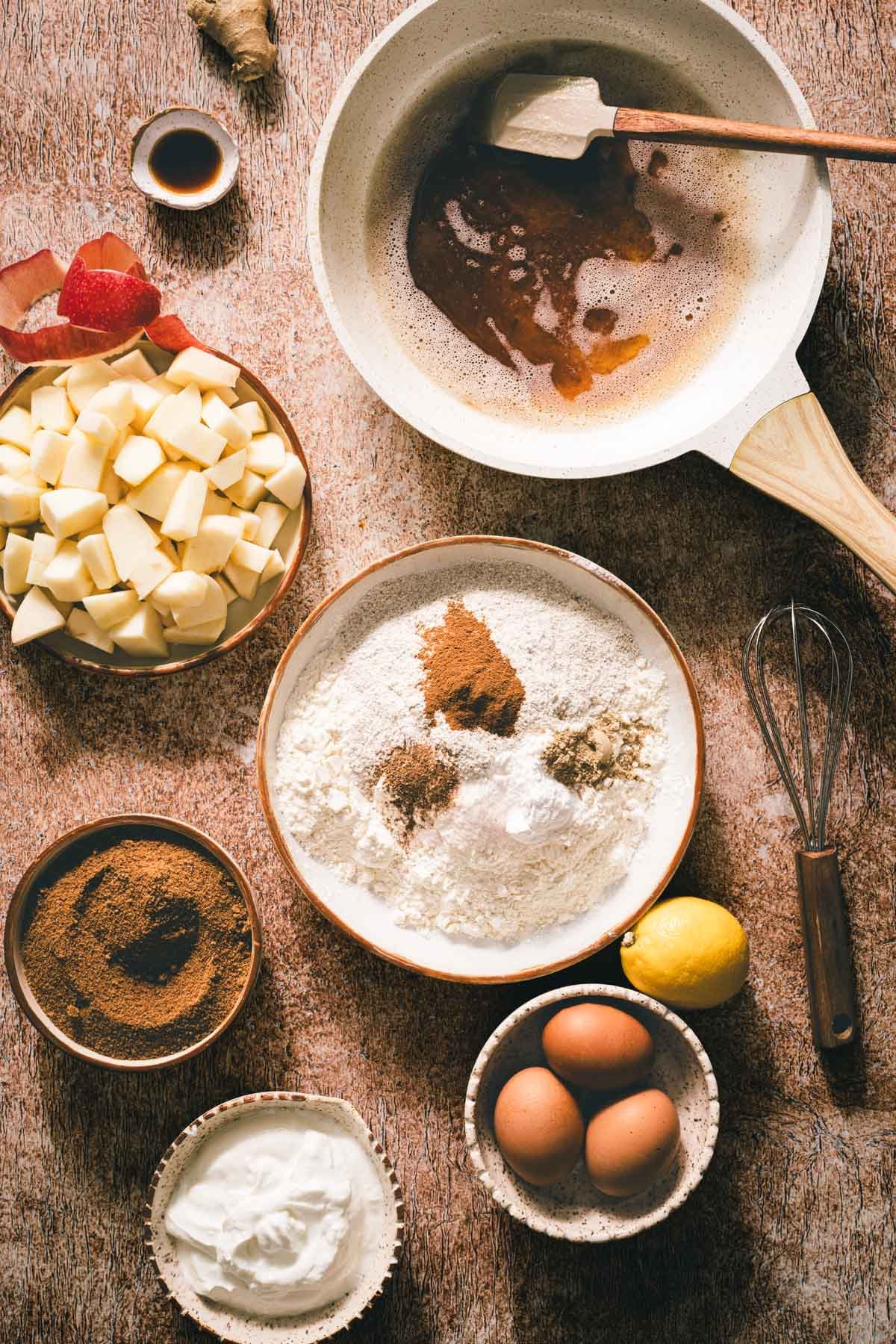 Browned butter in a skillet next to ingredients for apple cake: diced apples, spiced flour, sugar, yogurt, and eggs.