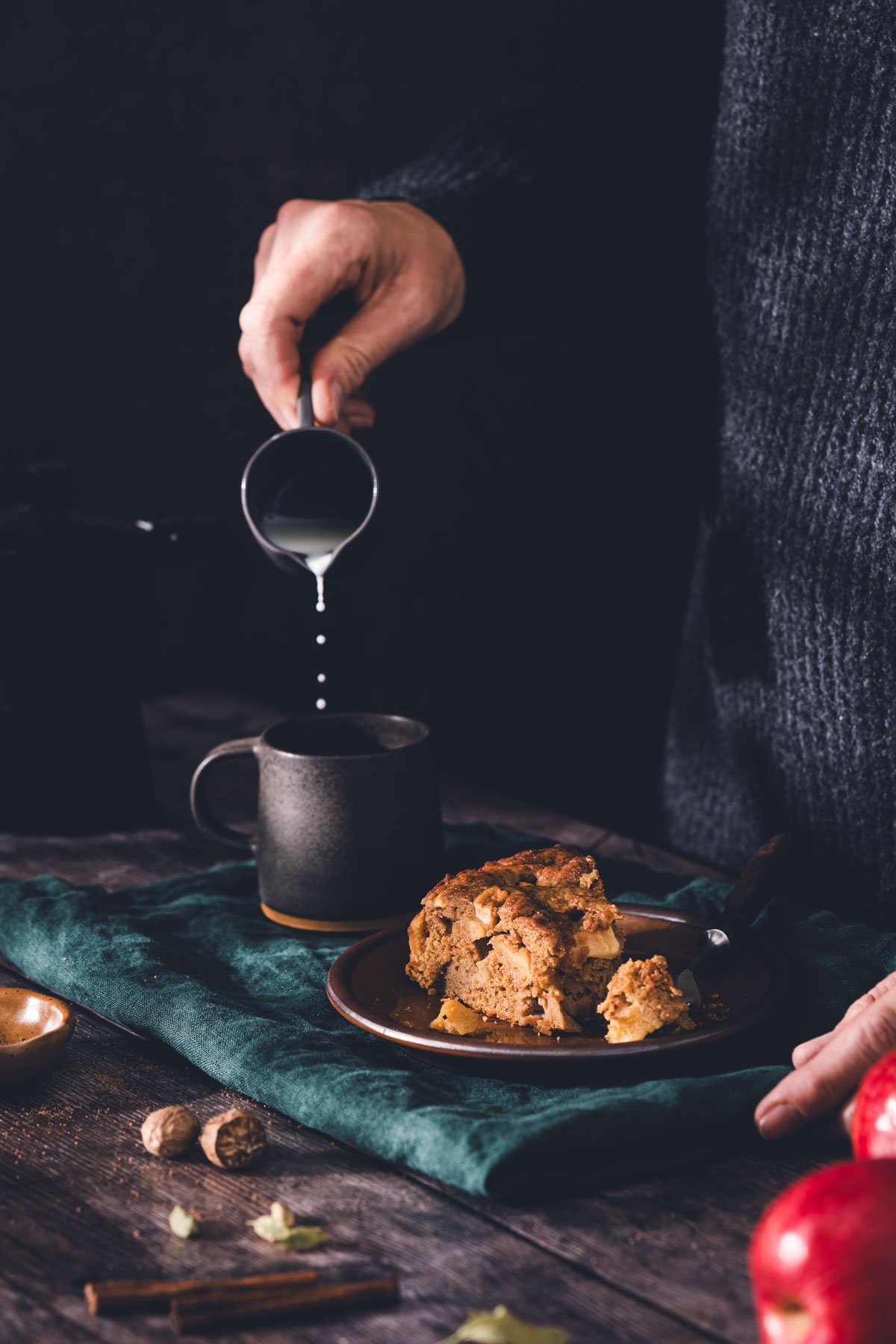 Hand pouring cream into a mug of coffee beside a slice of brown butter apple cake, with red apples in the foreground.