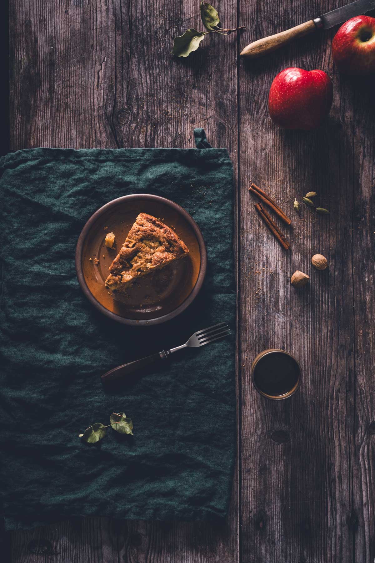 Top-down flat lay of a single brown butter apple cake slice on a brown plate, with fresh apples and whole spices on wood.