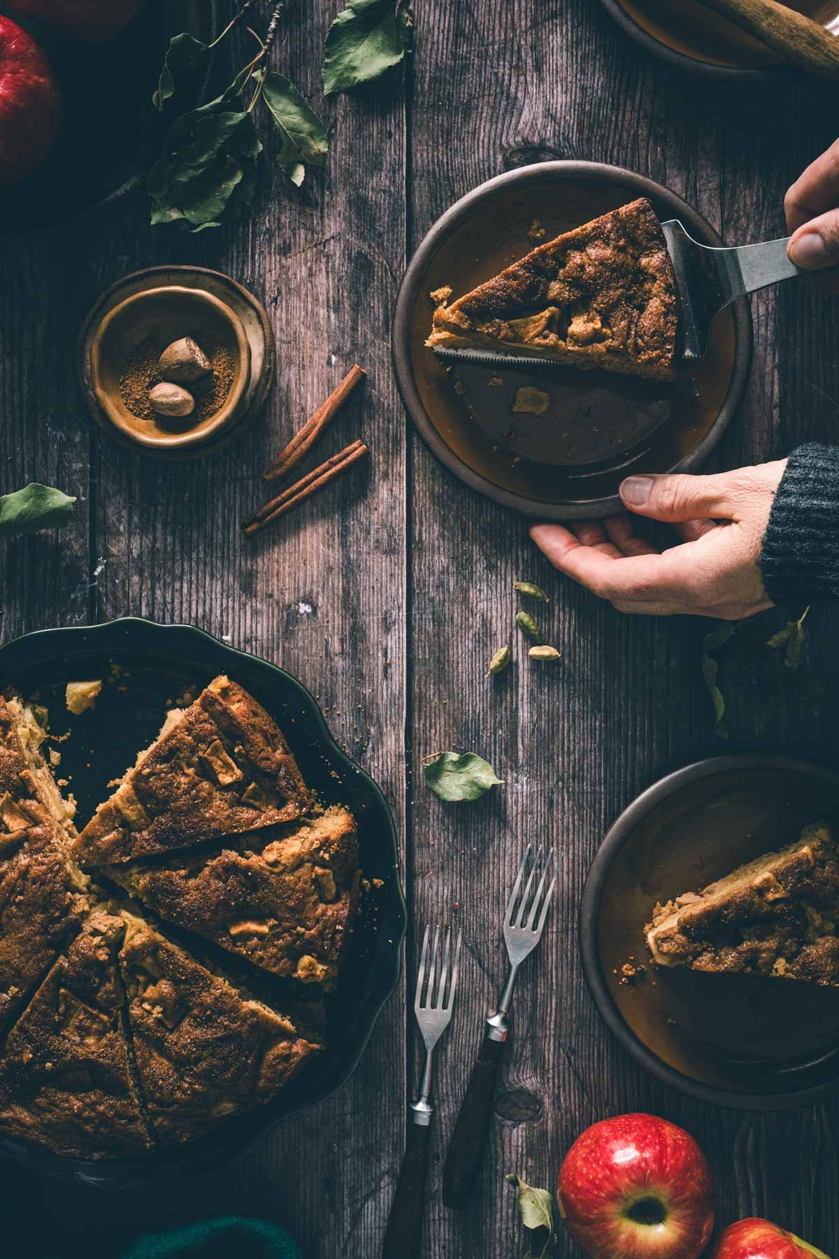 A hand serves a slice of brown butter apple cake onto a plate with a cake server, next to the sliced cake.