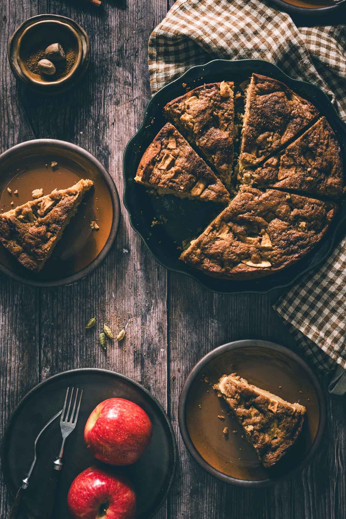 Top view of a whole caramelized apple cake in a green dish with a brown gingham towel, forks, and cinnamon sticks.