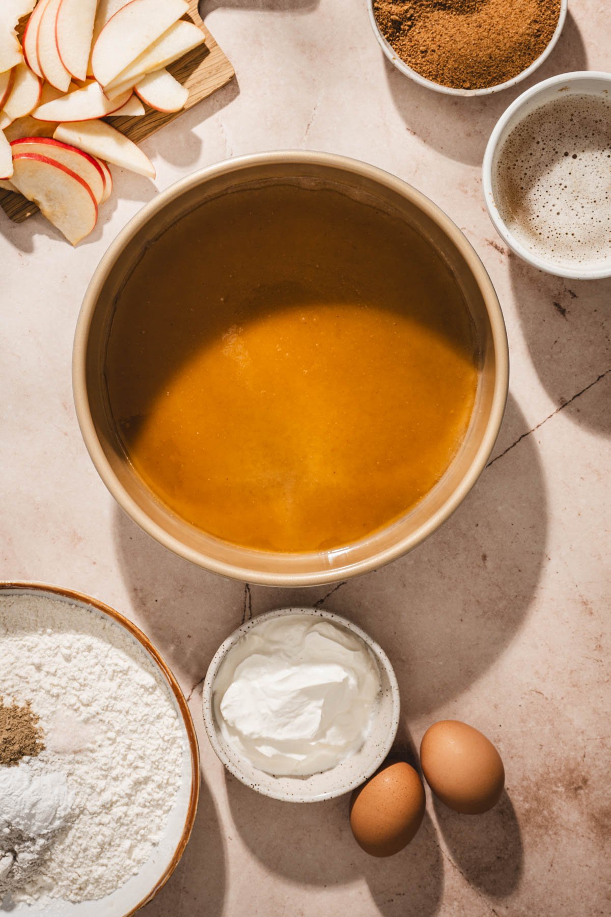 Golden caramel poured into the bottom of a gold cake pan, with apple slices, brown sugar, flour, yogurt, and eggs nearby.