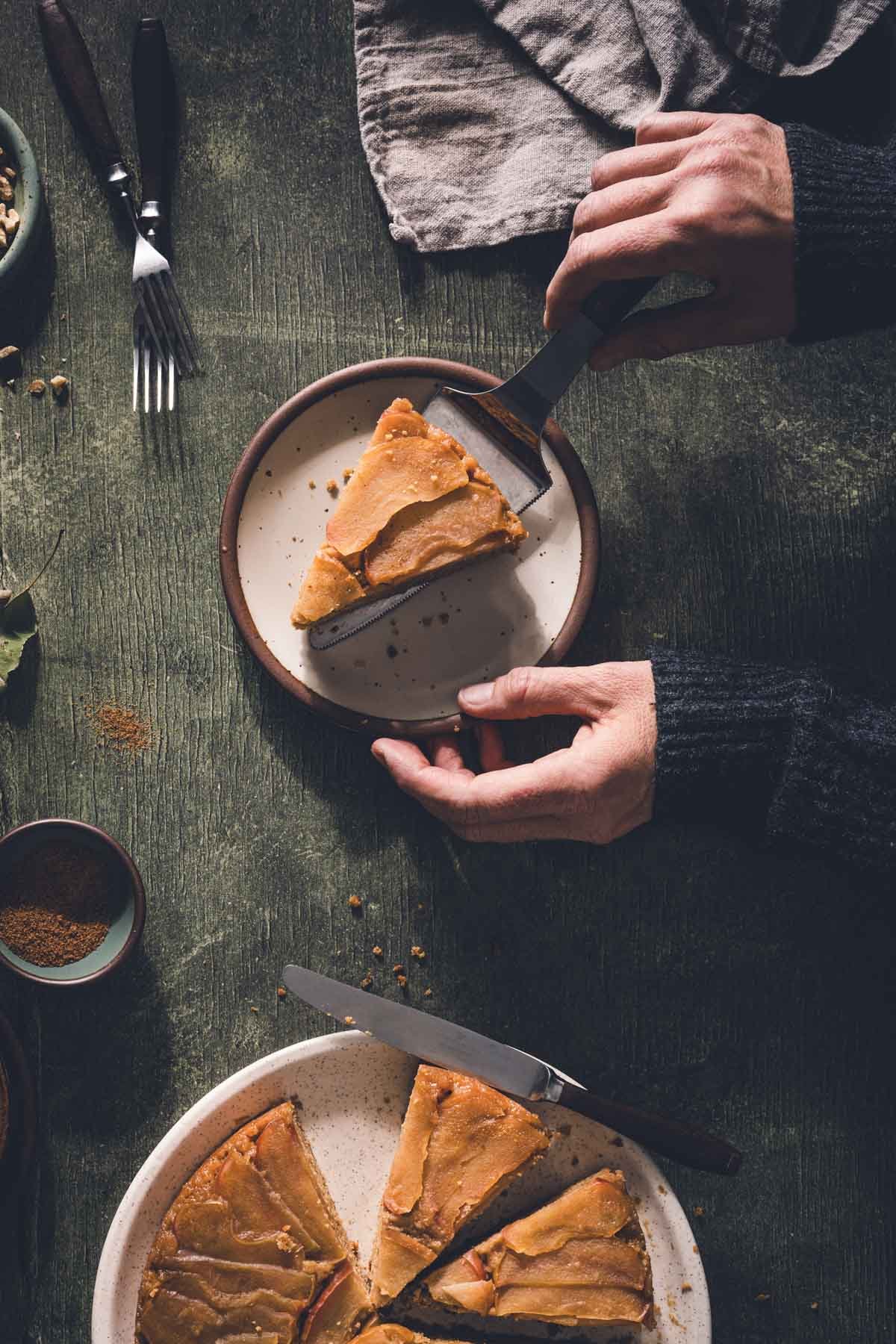 Hands using a cake server to place a slice of apple upside down cake onto a speckled plate.