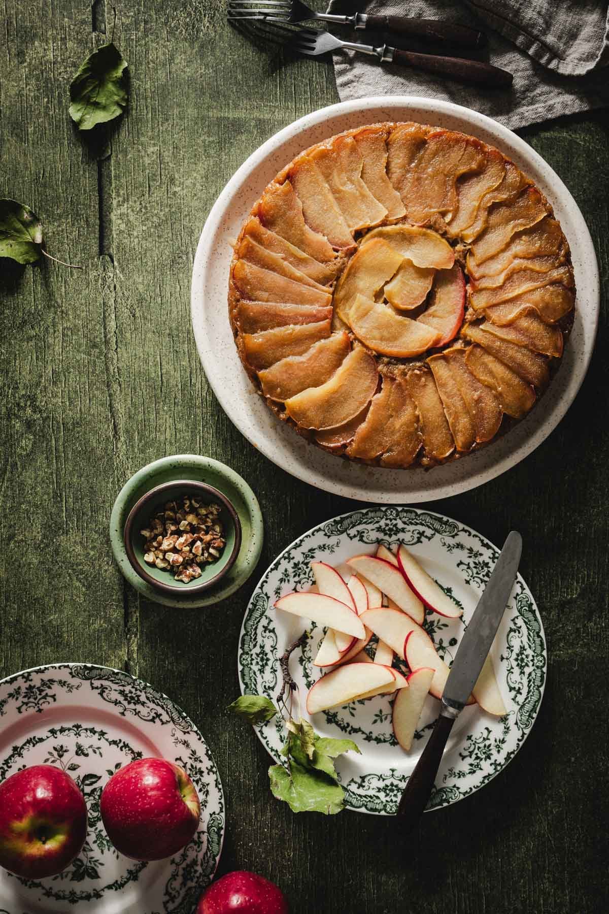 Whole apple upside down cake beside chopped walnuts, fresh apple slices, and red apples on vintage green plates.
