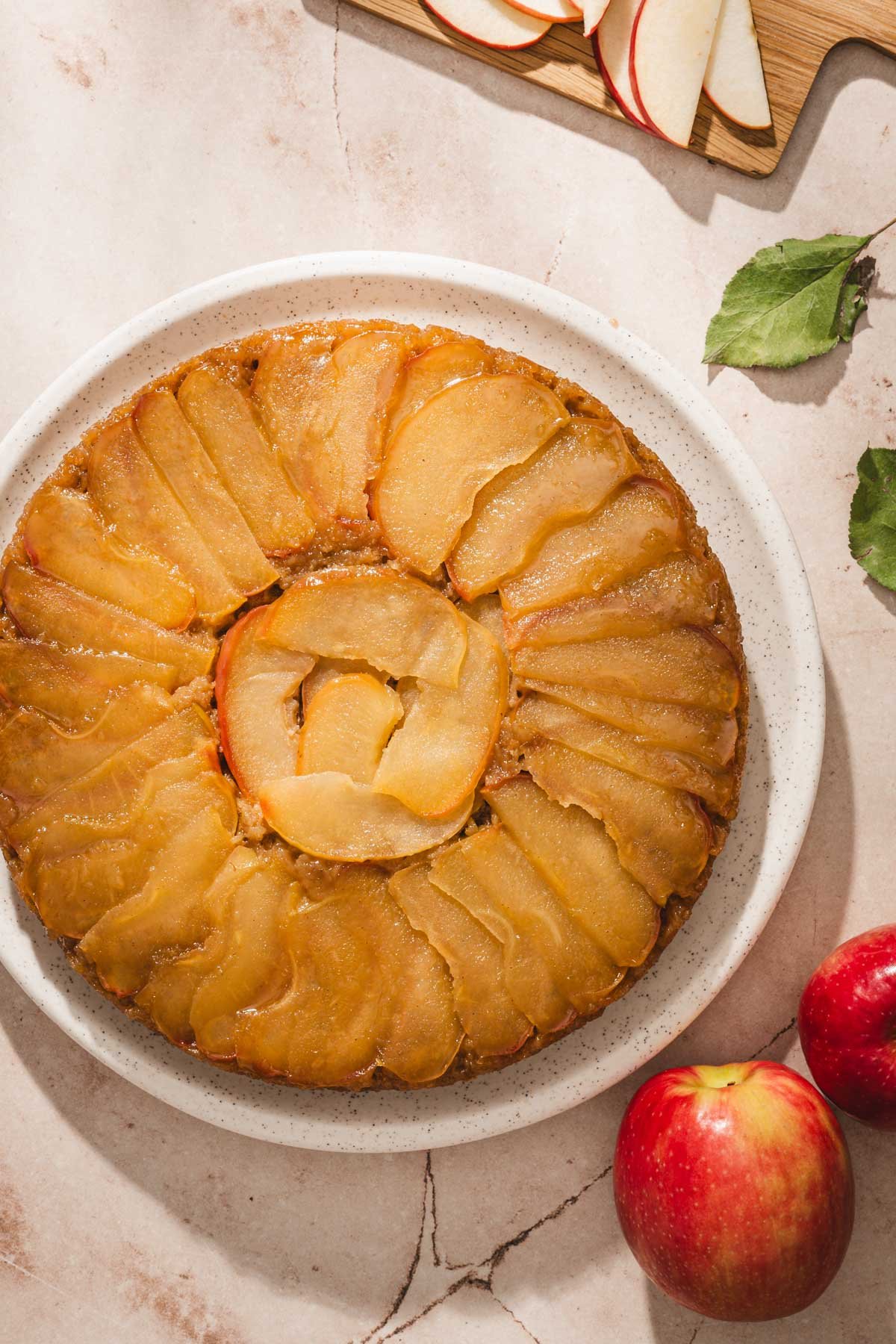 Whole apple upside down cake with a caramelized apple spiral on a speckled platter, with red apples on a light stone surface.