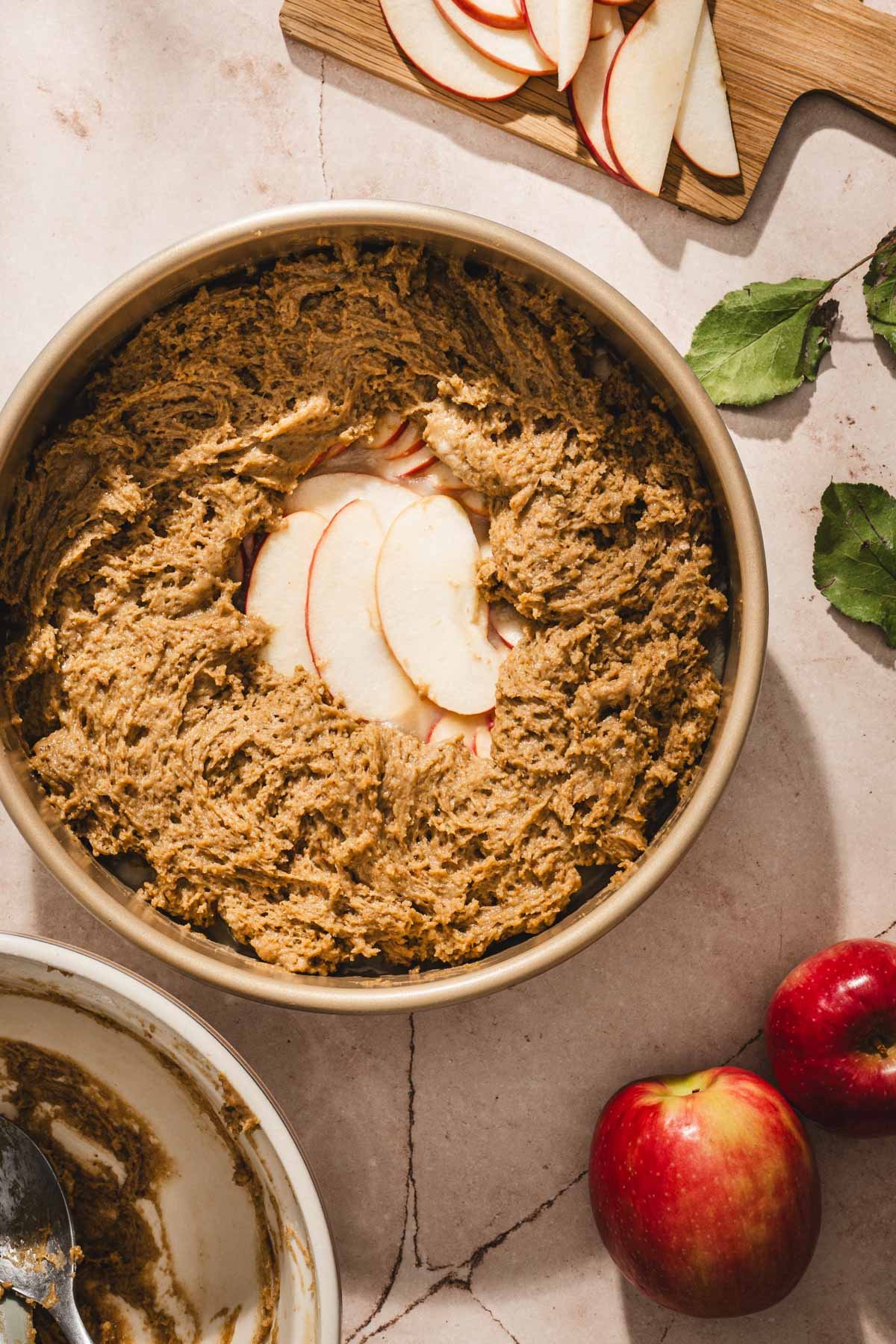 Cake batter spread over arranged apple slices in a gold cake pan, with a mixing bowl and fresh apples nearby.