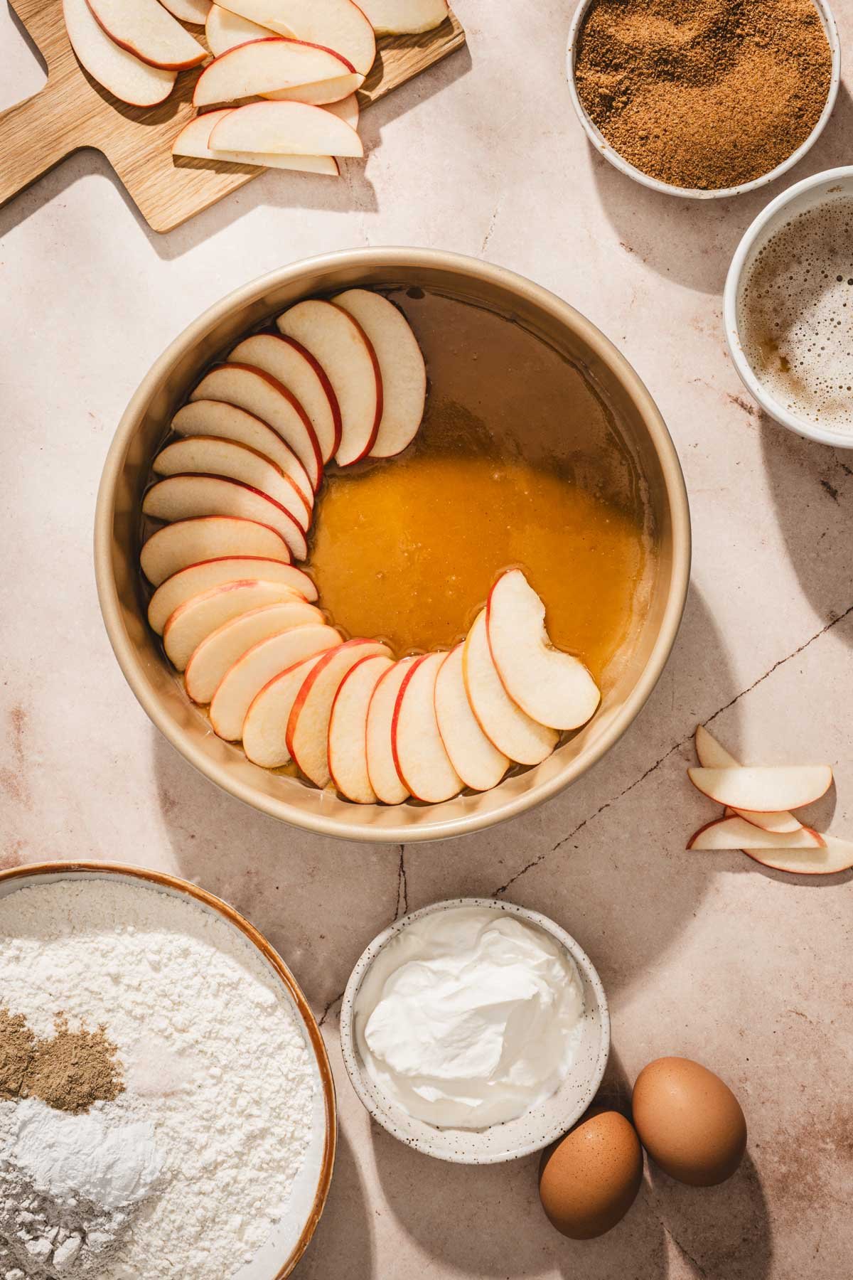 Apple slices being arranged in a circle over golden caramel in a cake pan, with flour, eggs, and yogurt nearby.