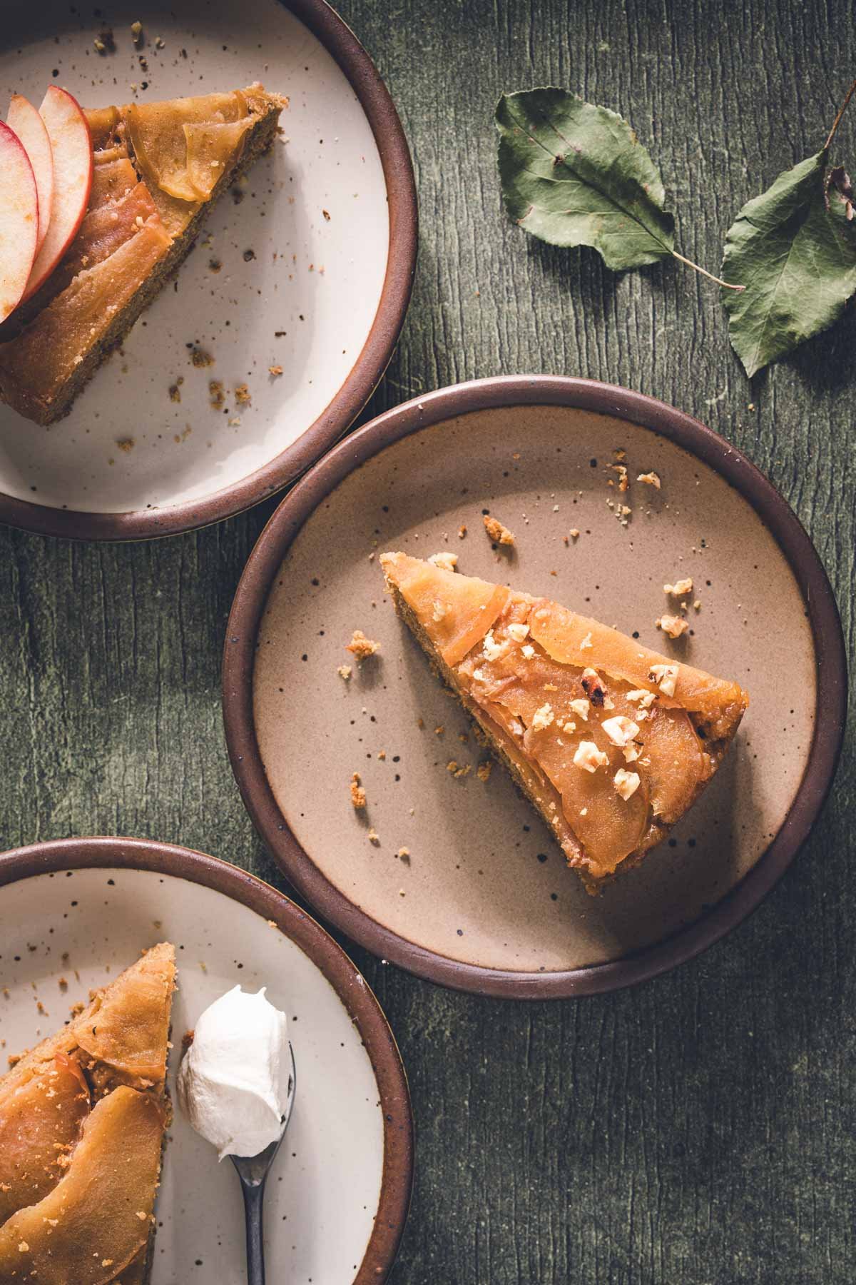 Three slices of apple upside down cake on speckled plates, one with chopped walnuts and one with whipped cream.