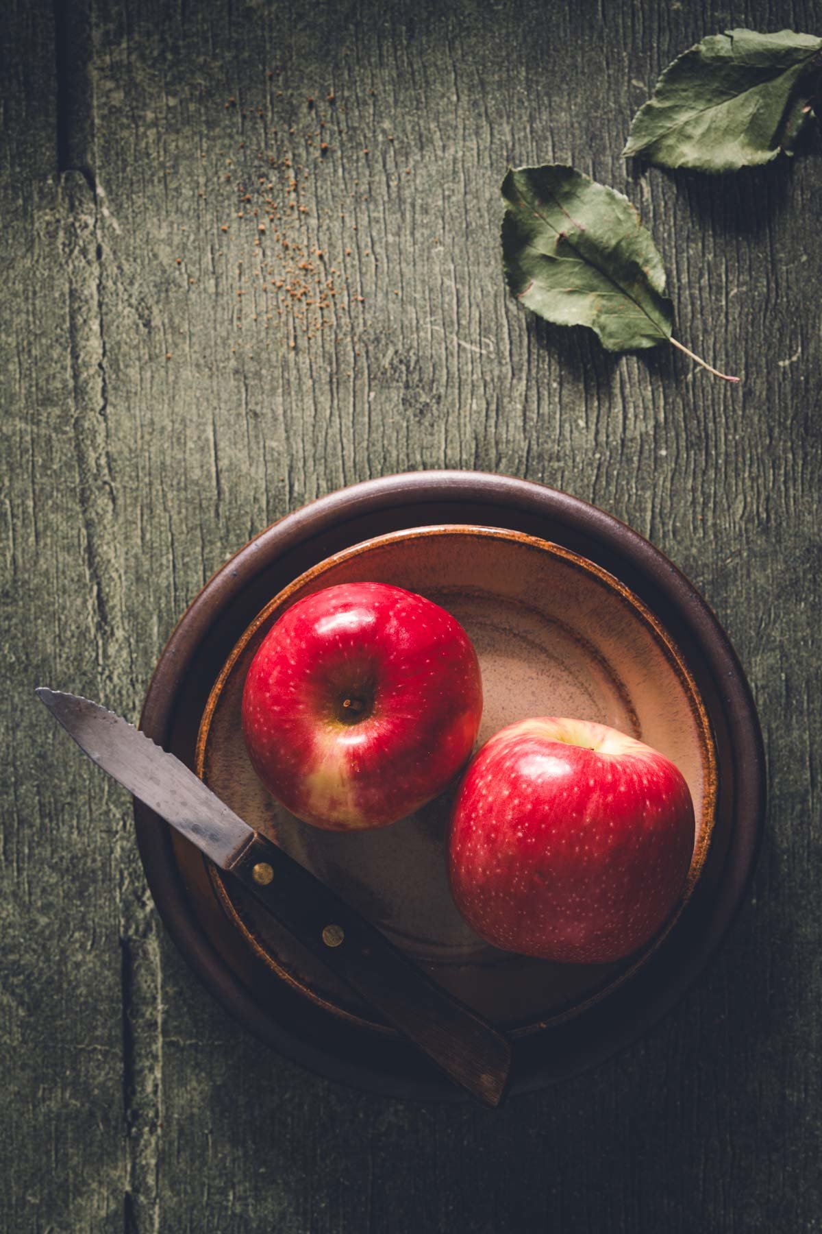 Two red apples on rustic stacked ceramic plates with a paring knife and dried apple leaves.