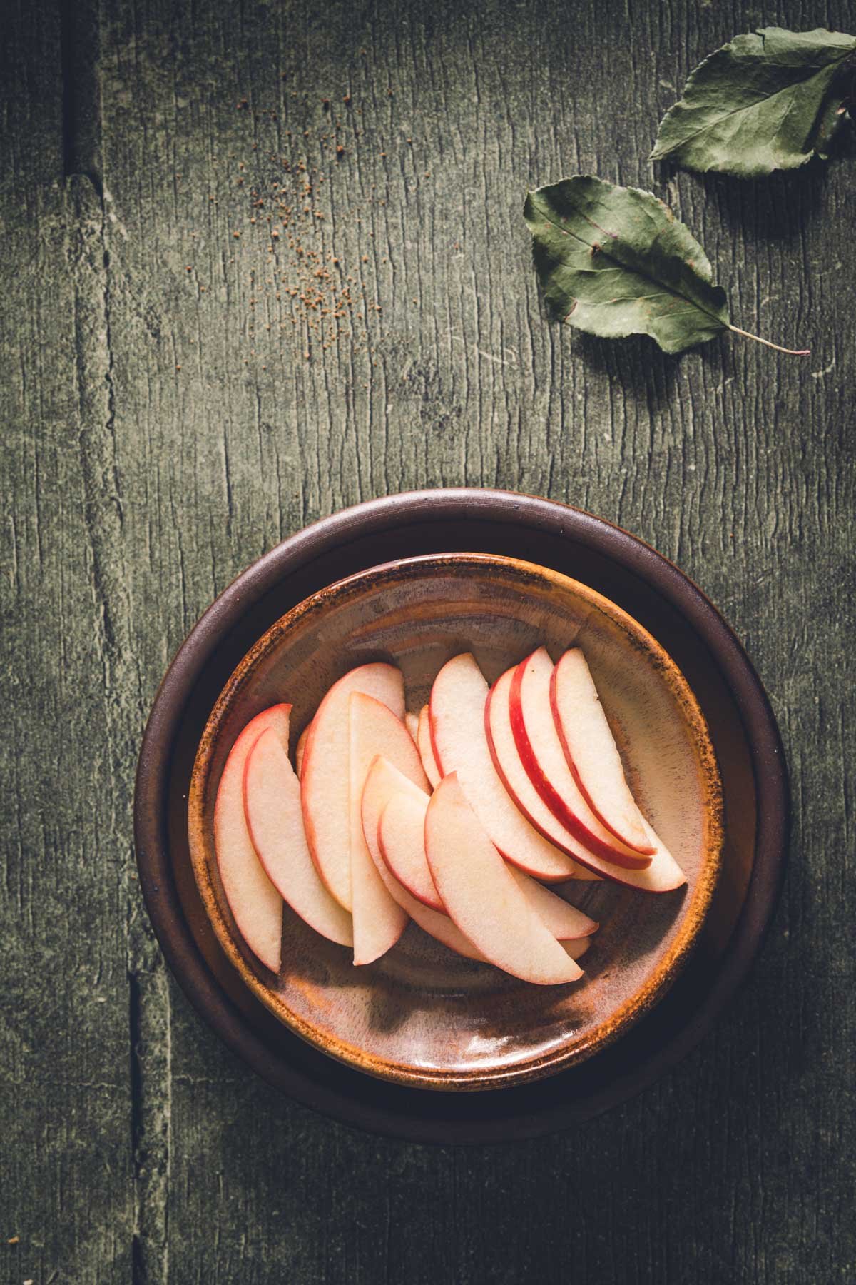 Fresh red apple slices fanned out on a rustic brown glazed ceramic plate on dark green wood.