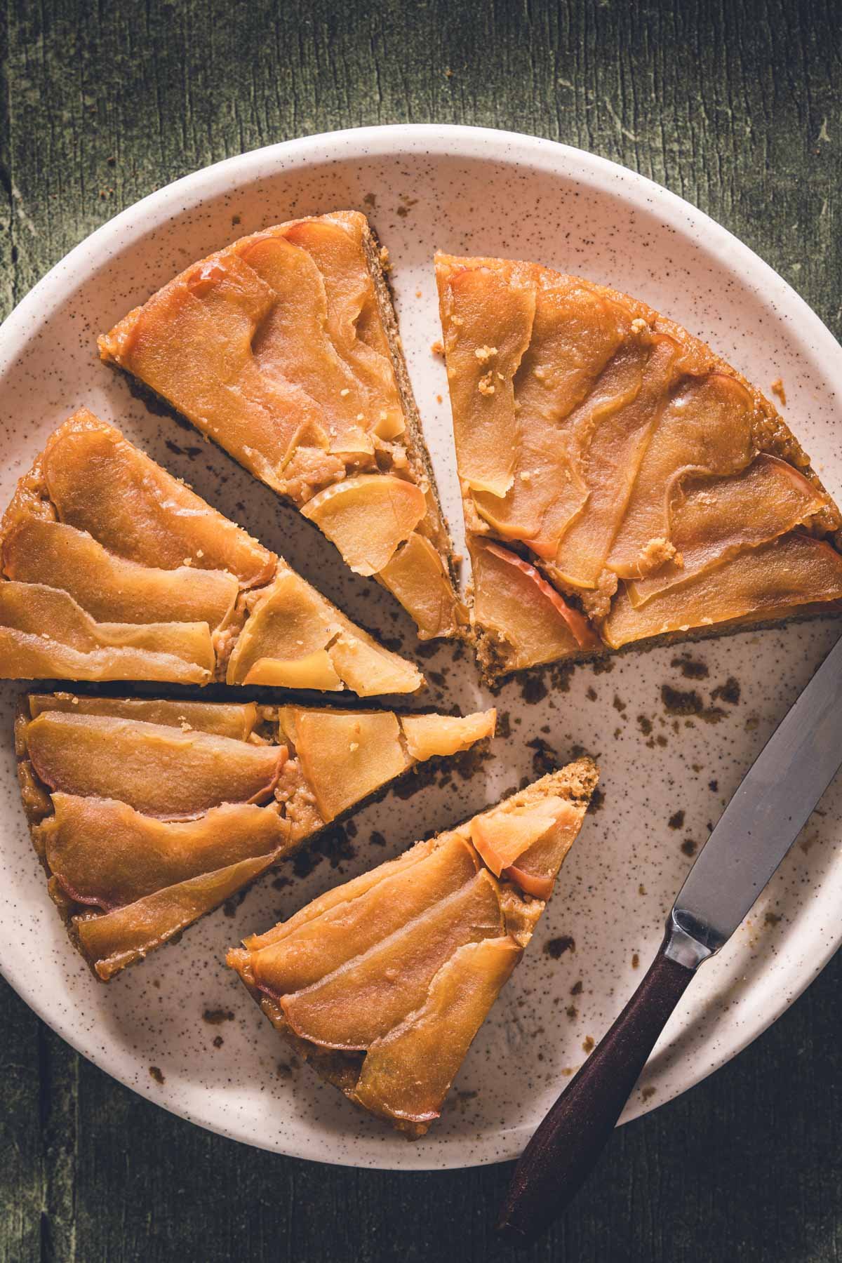 Close-up of apple upside down cake cut into wedges on a speckled platter with a wooden-handled knife.