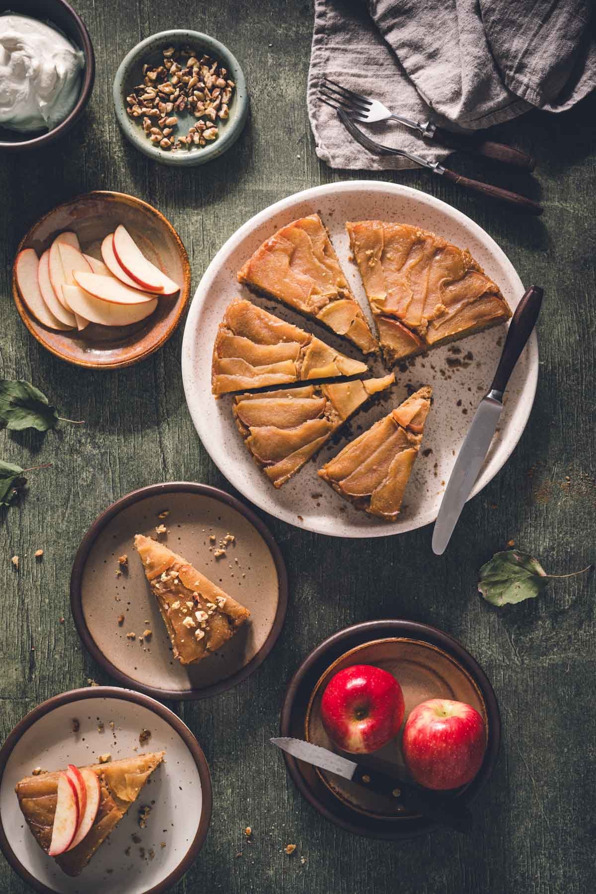 Overhead table spread of sliced apple upside down cake with walnuts, whipped cream, apple slices, and forks.