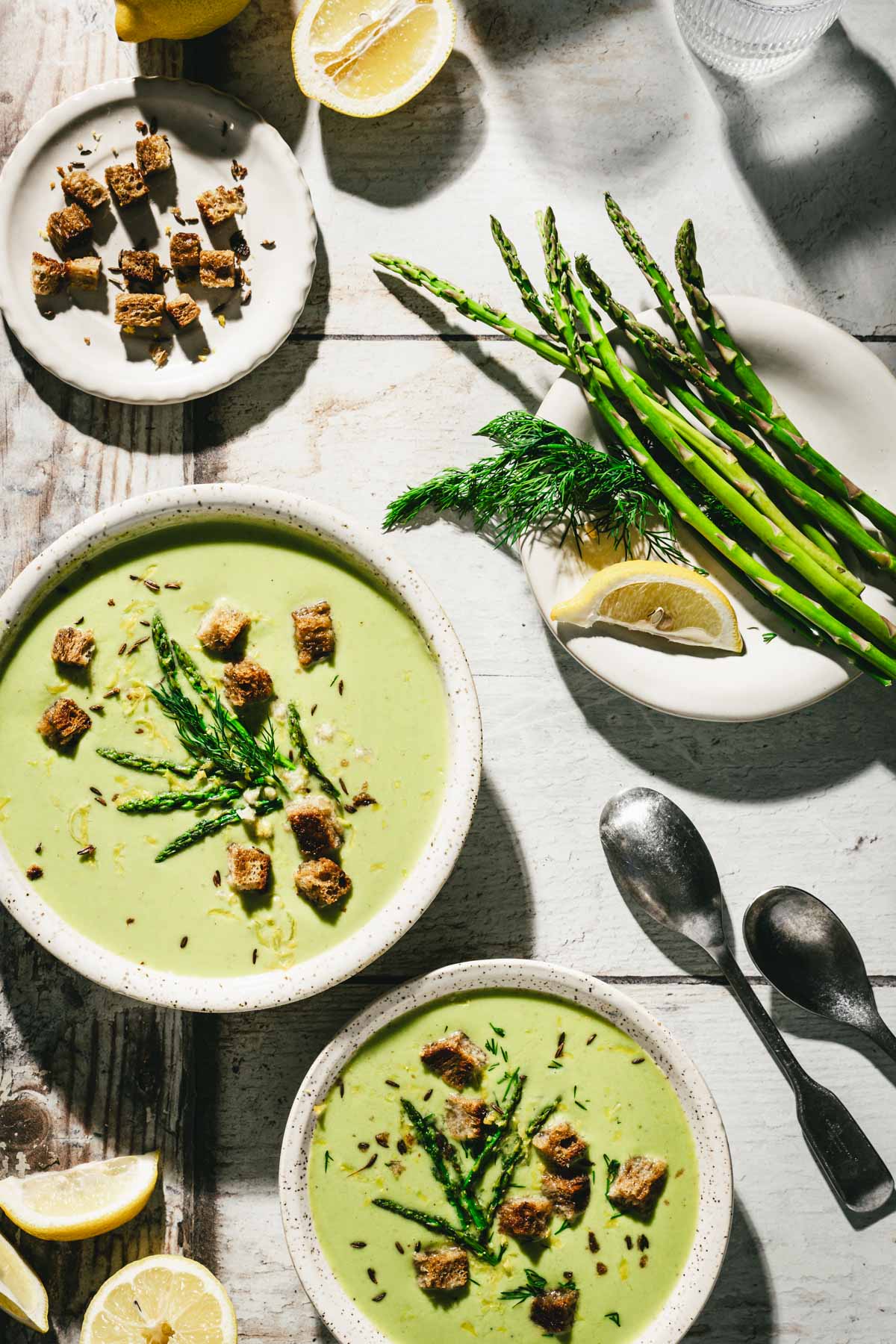 various serving bowls with soup, spoons, lemon wedges, dish with fresh dill and asparagus