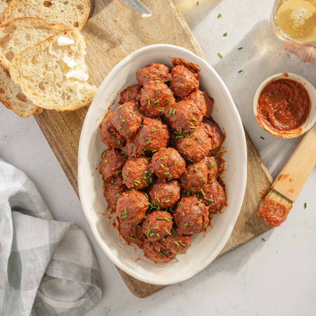 BBQ Meatballs on a sesrving platter, slices of bread, two glasses, dish with bbq sauce and a sauce brush