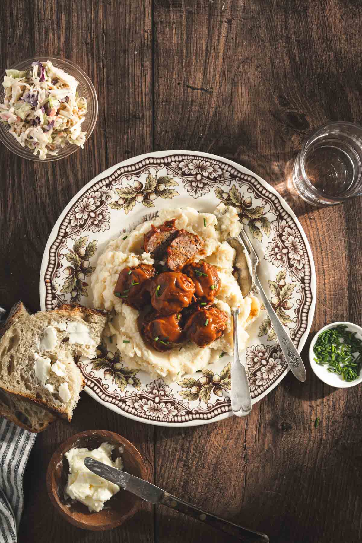 bbq meatballs on a serving plate with mashed potatoes, slice of bread with butter, glass of water, small dish with salad and chives