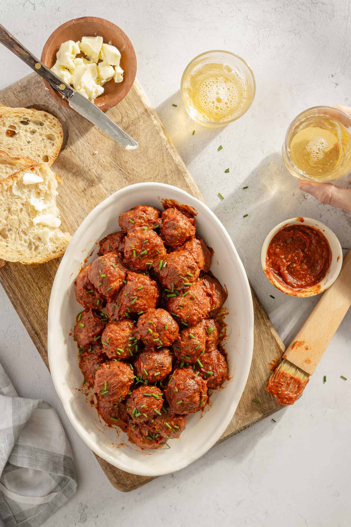 BBQ Meatballs on a sesrving platter, slices of bread, two glasses, dish with bbq sauce and a sauce brush