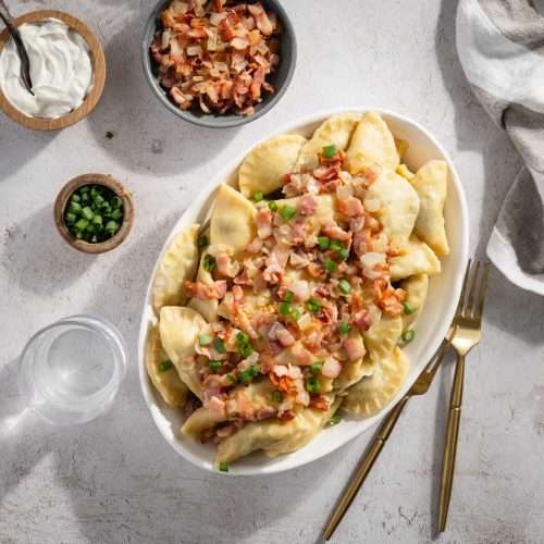 bowl with cooked pierogis with bacon and onion topping; serving utensils, small bowl with chives and bacon