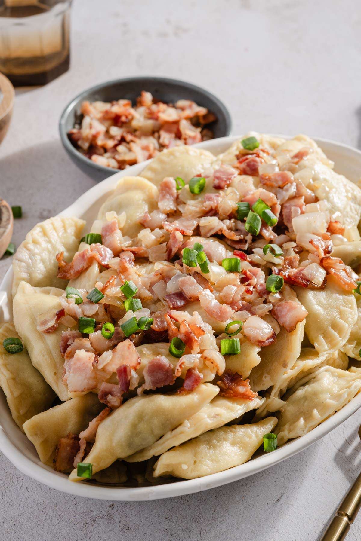 bowl with cooked pierogis with bacon and onion topping; serving utensils, small bowl with chives and bacon