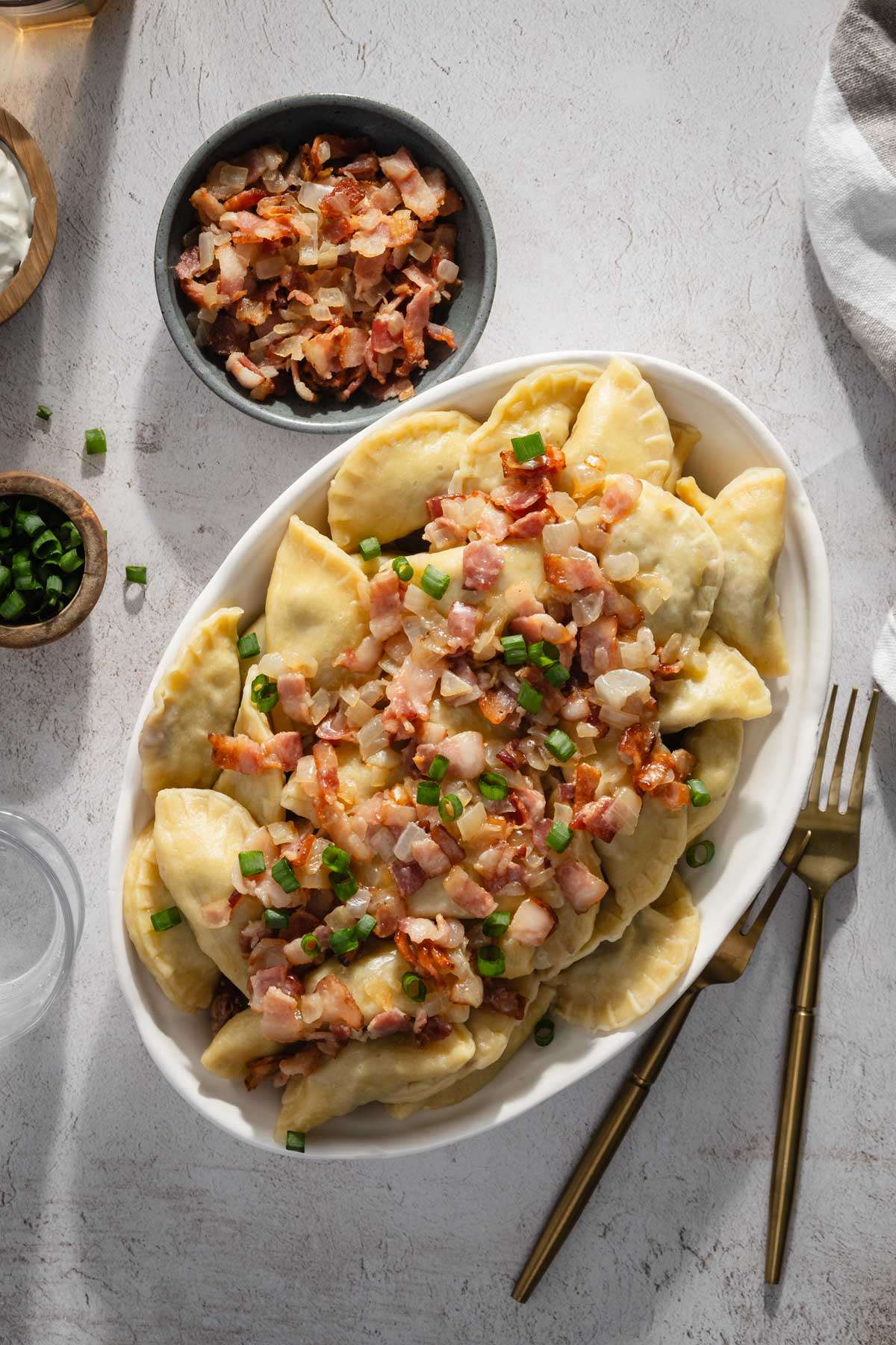 bowl with cooked pierogis with bacon and onion topping; serving utensils, small bowl with chives and bacon