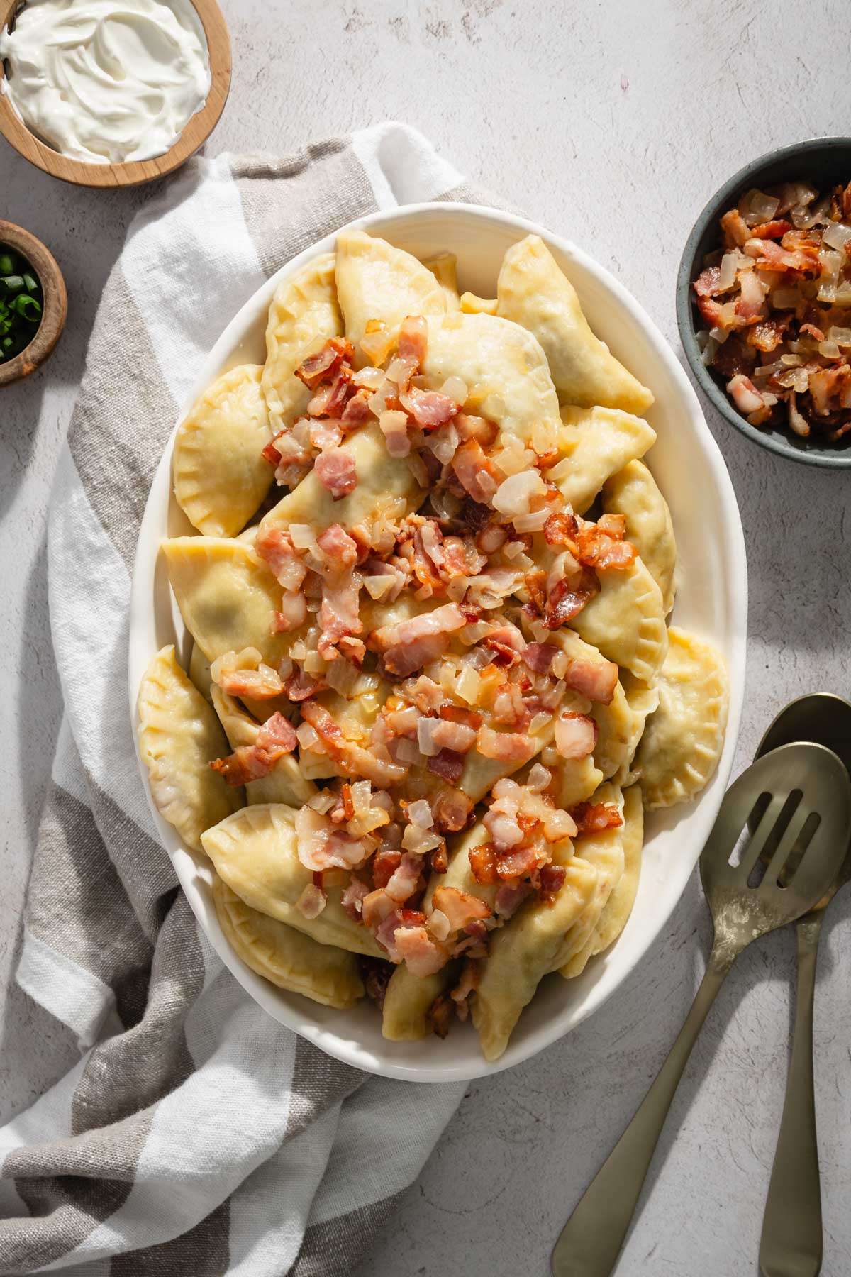 bowl with cooked pierogis with bacon and onion topping; serving utensils, small bowl with chives and bacon