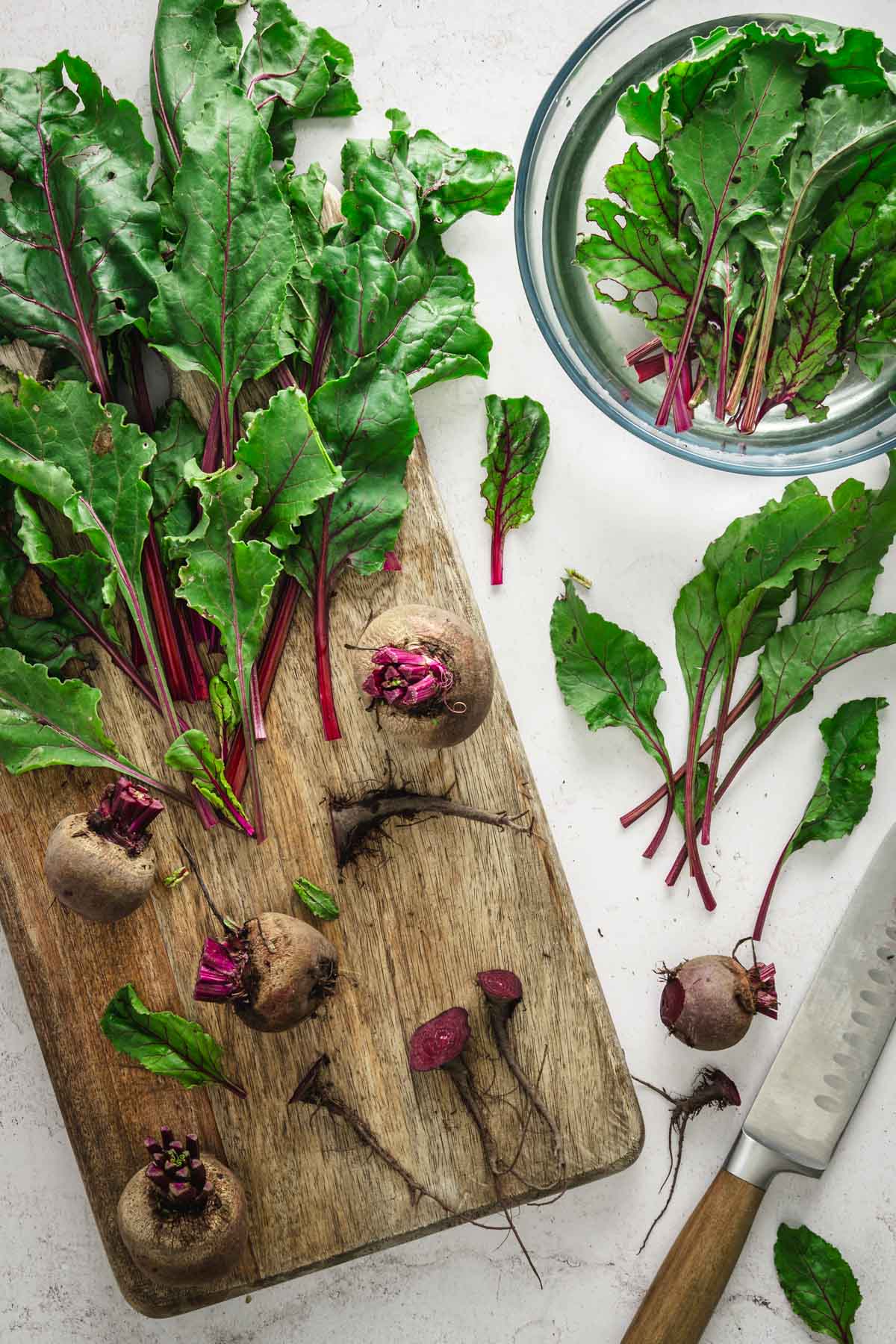 beet leaves on a wooden board, some in a glass bowl with water, knife