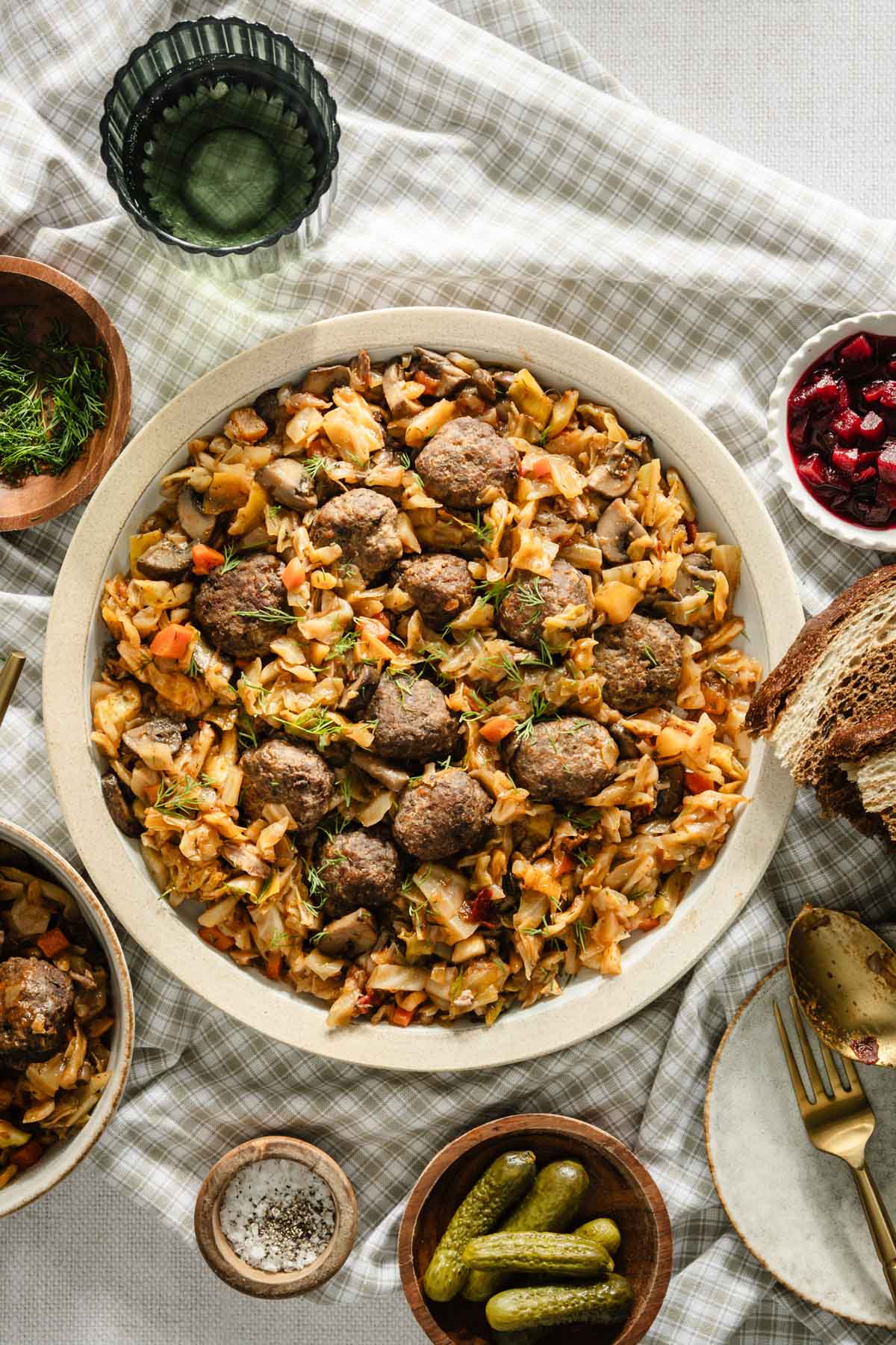 cooked smoked cabbage on a serving plate with meatballs, bread, pickles, fresh dill, pickled beets, plate with forks