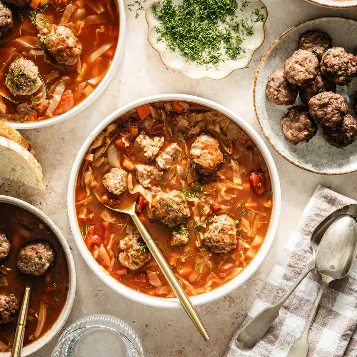 Cabbage soup served in bowls, meatballs on the side, bread knife, bread basket, dish with fresh dill