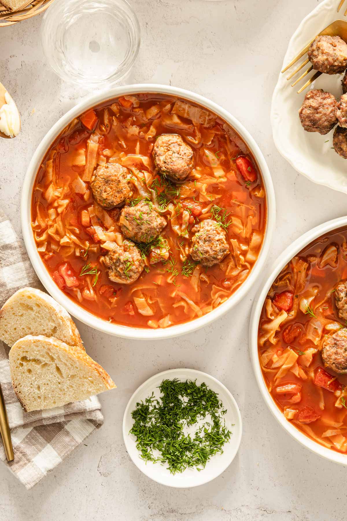 Cabbage soup served in bowls, meatballs on the side, bread knife, bread basket, utensils