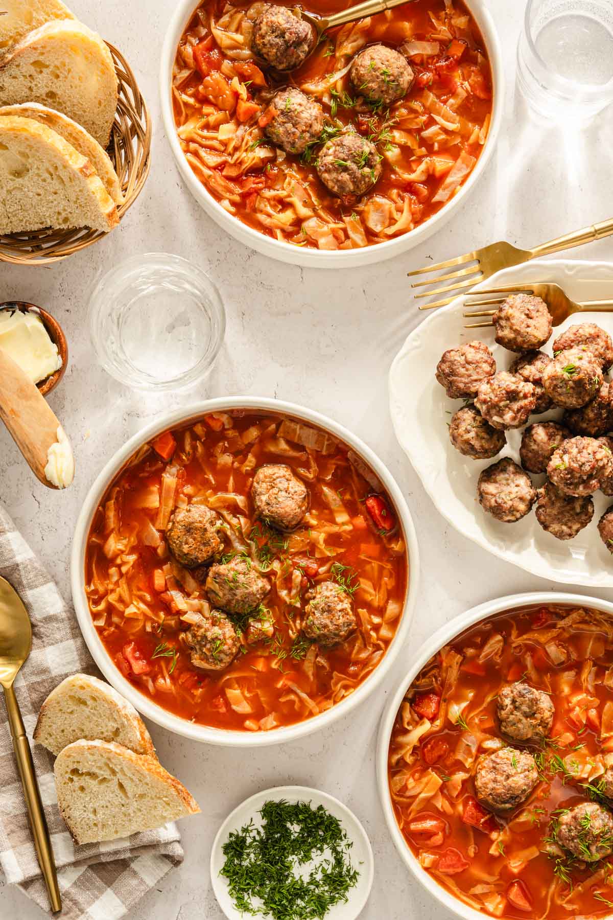 Cabbage soup served in bowls, meatballs on the side, bread knife, bread basket, utensils