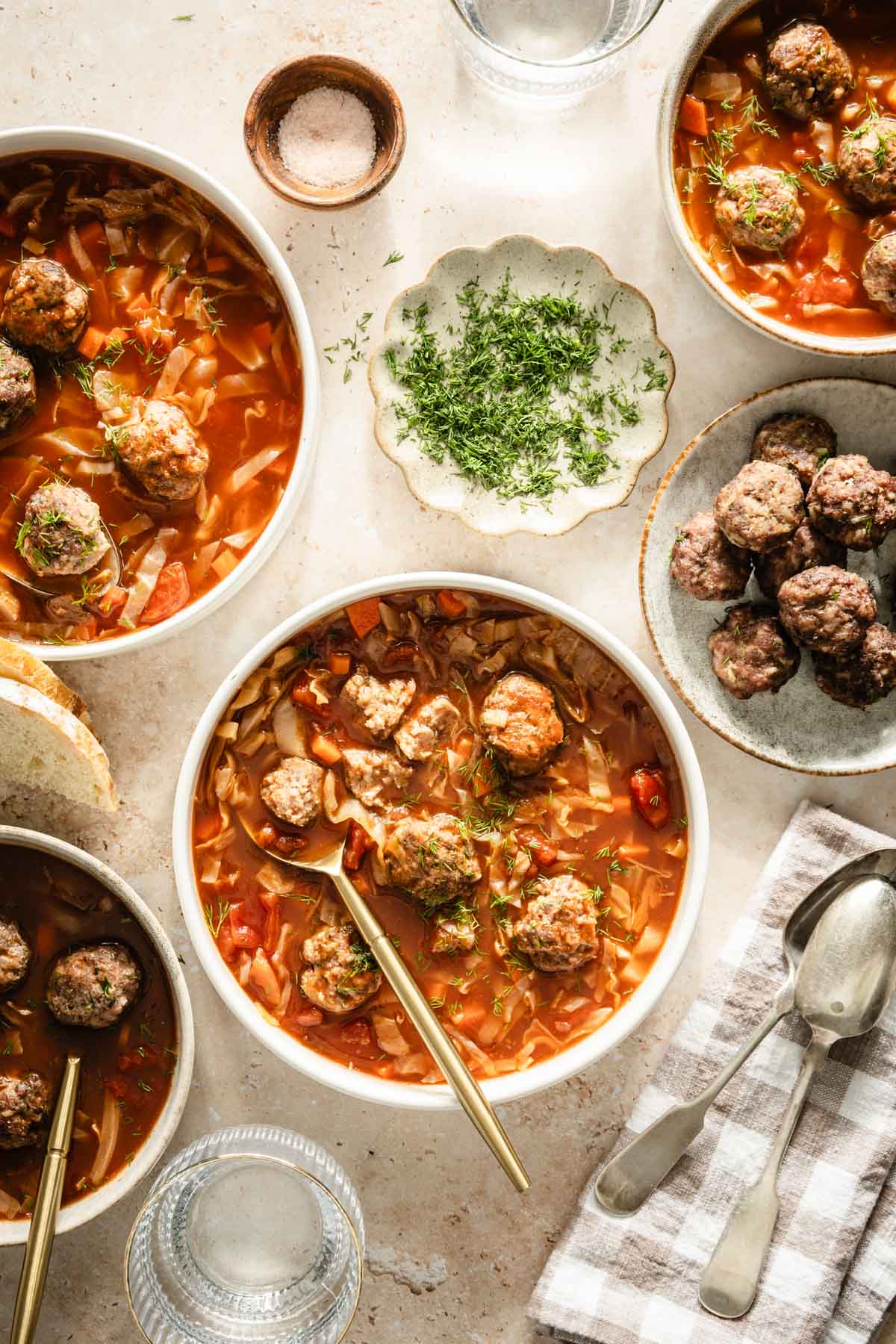 Cabbage soup served in bowls, meatballs on the side, bread knife, bread basket, dish with fresh dill, utensils, glass of water and napkin