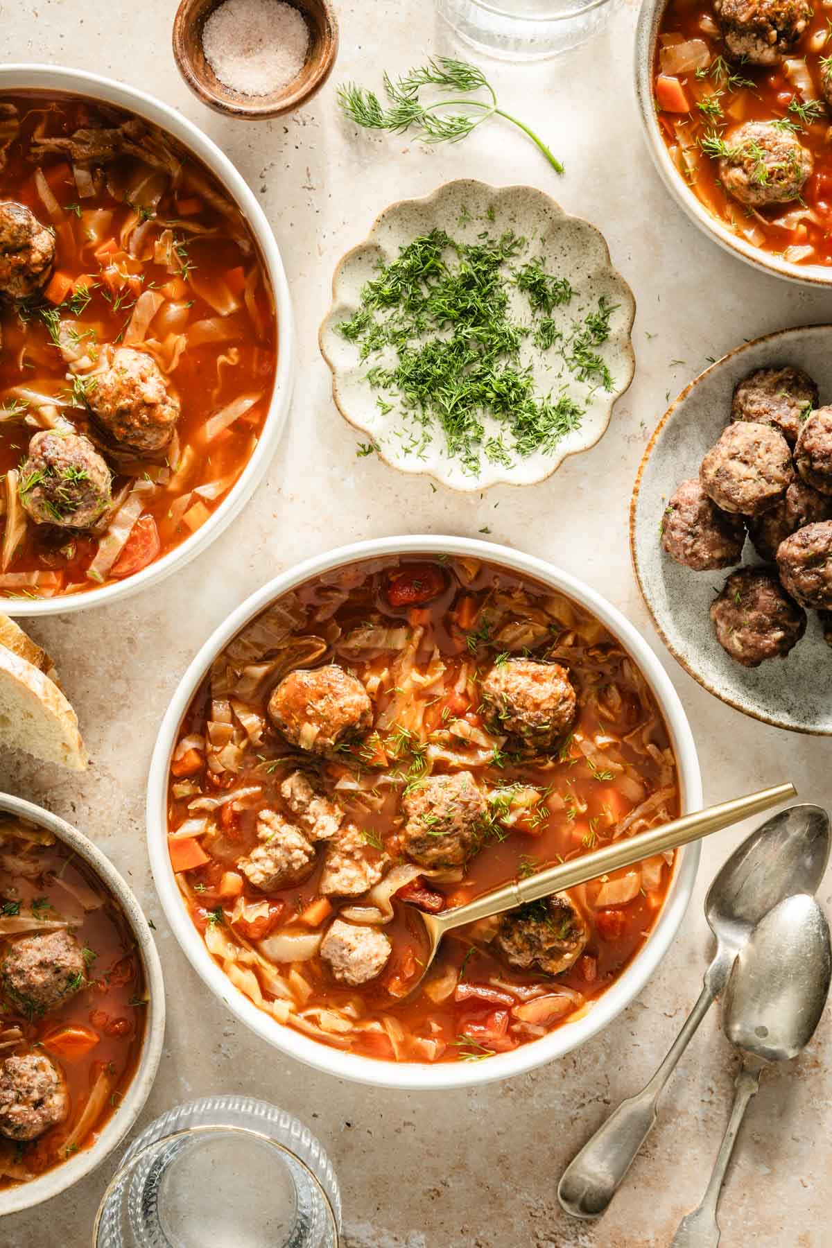 Cabbage soup served in bowls, meatballs on the side, bread knife, bread basket, dish with fresh dill