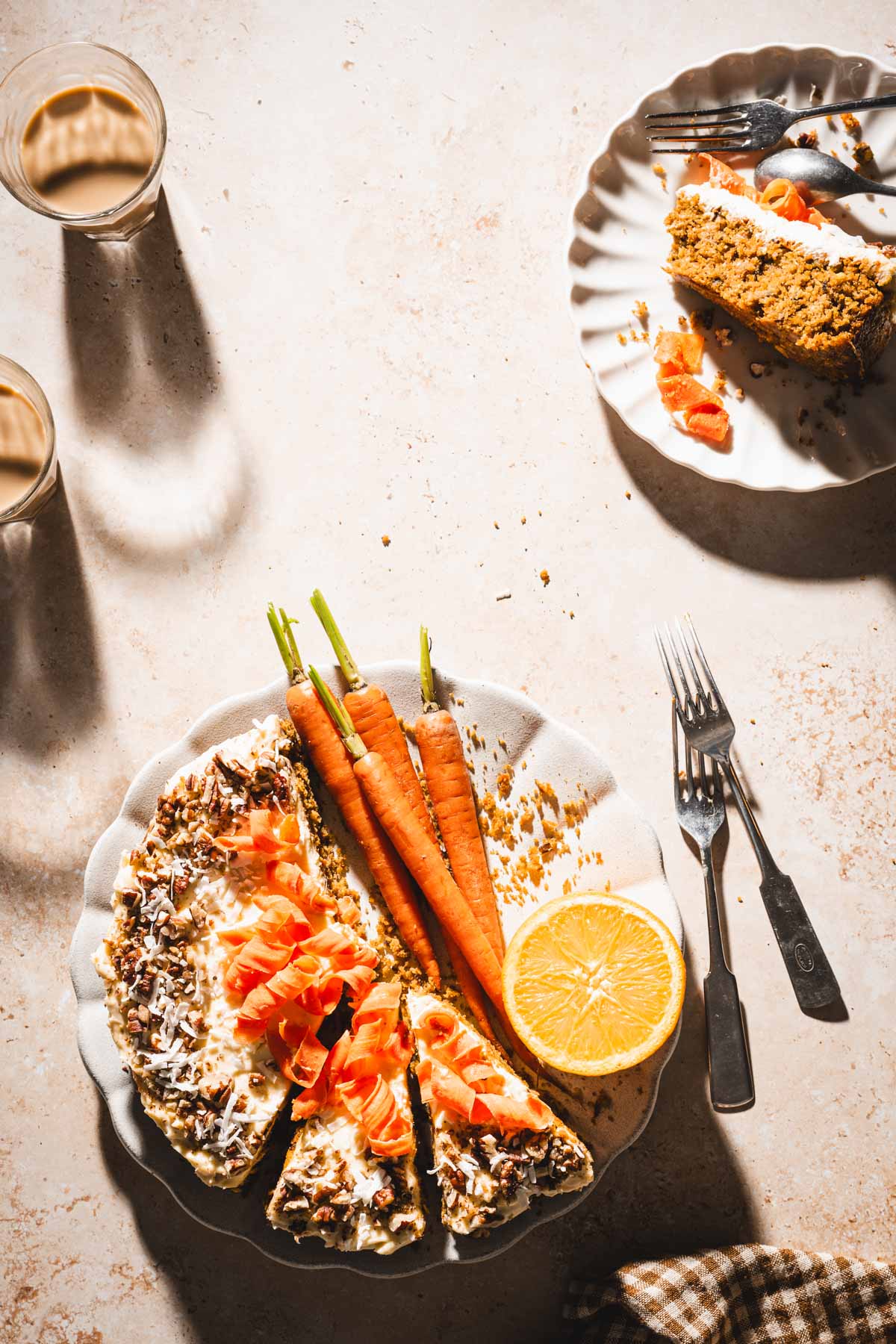 cake on a serving plate, fresh carrots, two glasses with coffee, two forks, plate with a slice of cake
