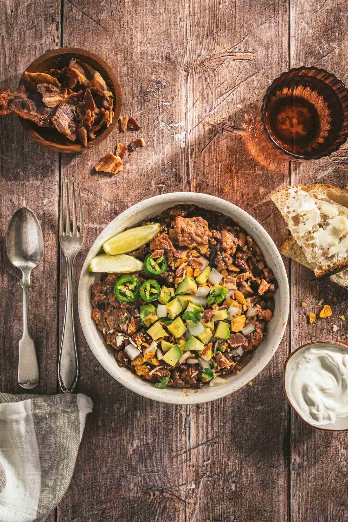 chili in a serving bowl garnished with diced avocado, limes, jalapenos, utensils on the left, glass of water, bowl with bacon, sour cream, two slices of bread, napkin