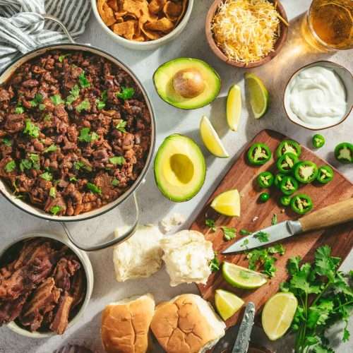 table scene with pan filled with chili, cut up avocado, cutting board with knife, limes, cilantro, bread buns, little bowls with sides: cheese, chips, sour cream, bacon