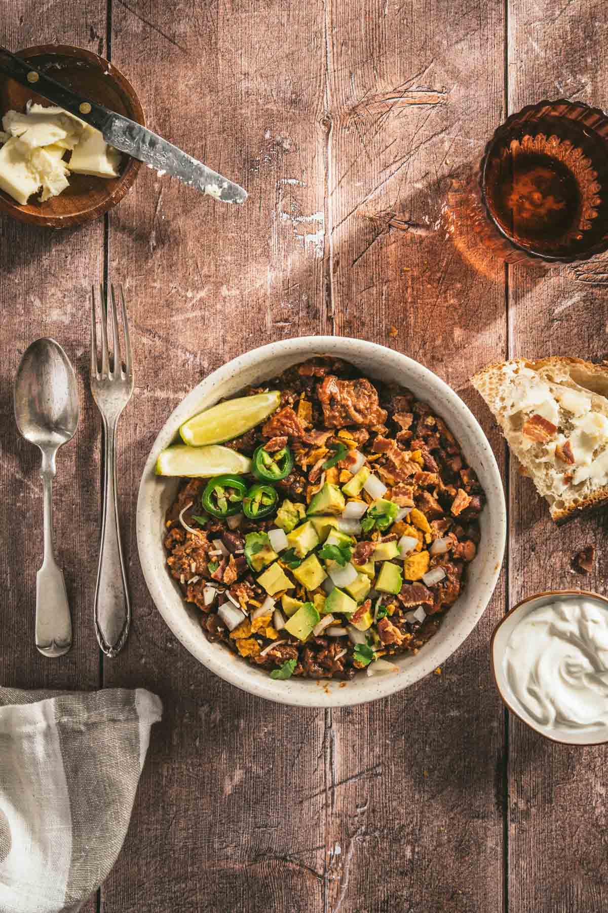 chili in a serving bowl garnished with diced avocado, limes, jalapenos, utensils on the left, glass of water, bowl with bacon, sour cream, two slices of bread, napkin