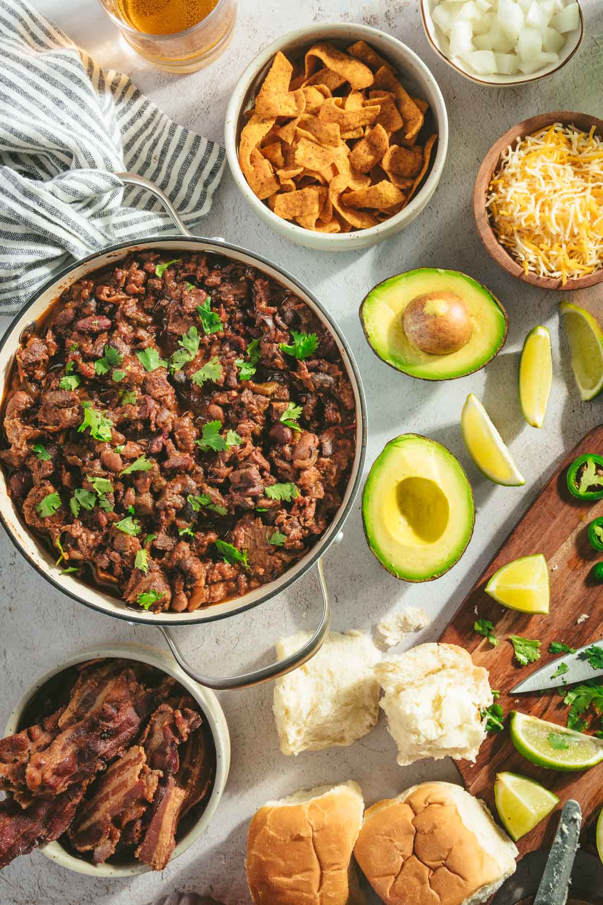 table scene with pan filled with chili, cut up avocado, cutting board with knife, limes, cilantro, bread buns, little bowls with sides: cheese, chips, sour cream, bacon