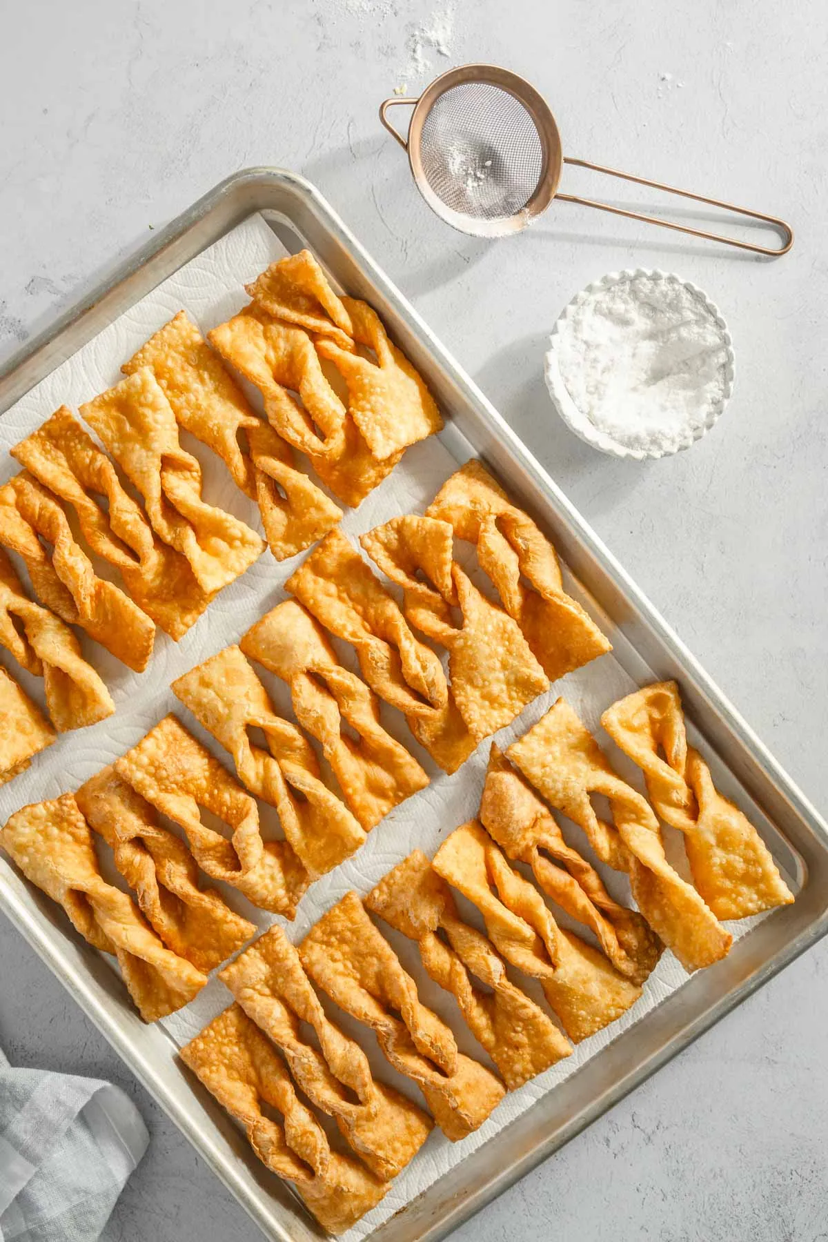 cooked chrusciki on a baking sheet, small bowl with powdered sugar, spoon and sieve