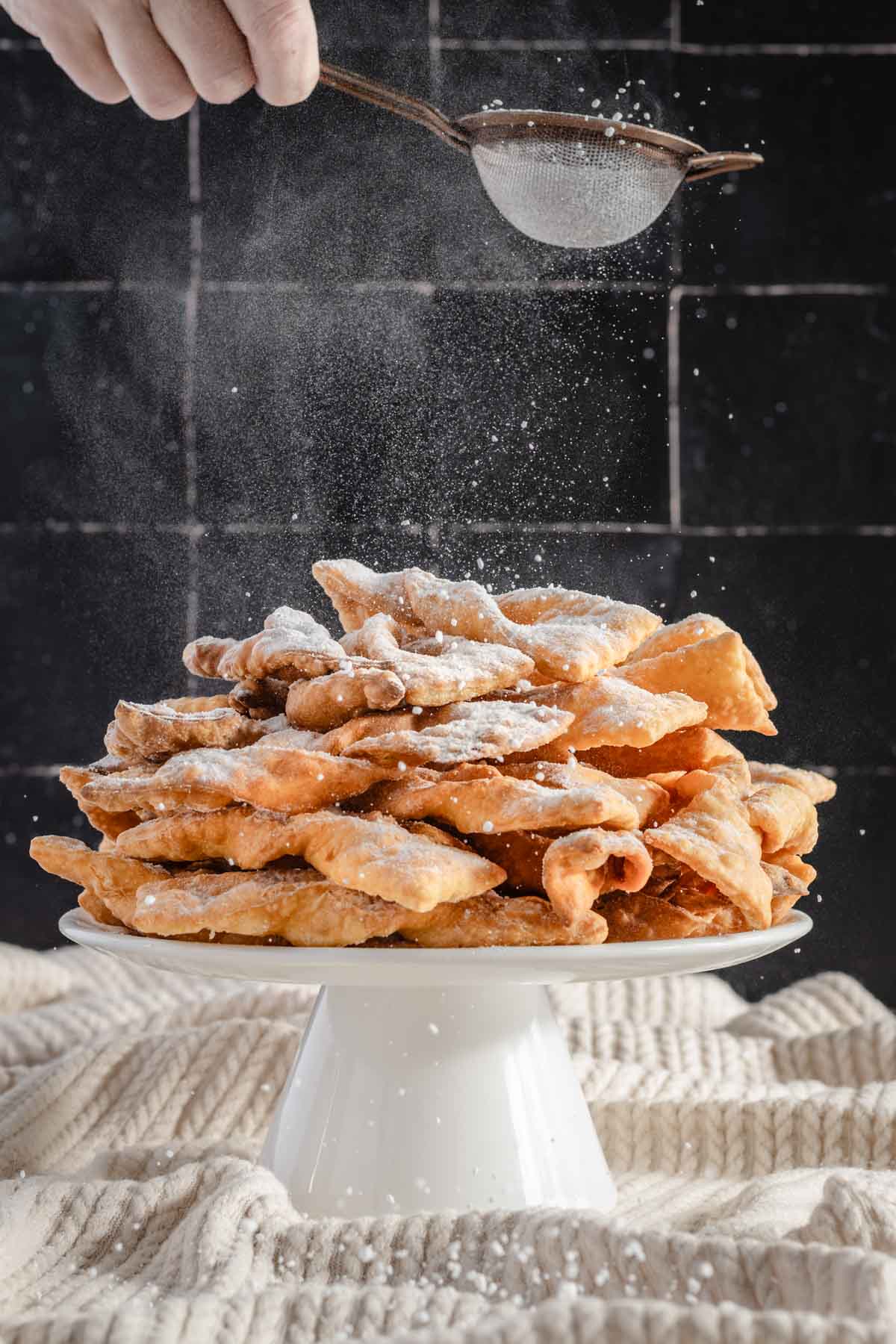 chrusciki on a cake stand, hand in frame drizzling powdered sugar