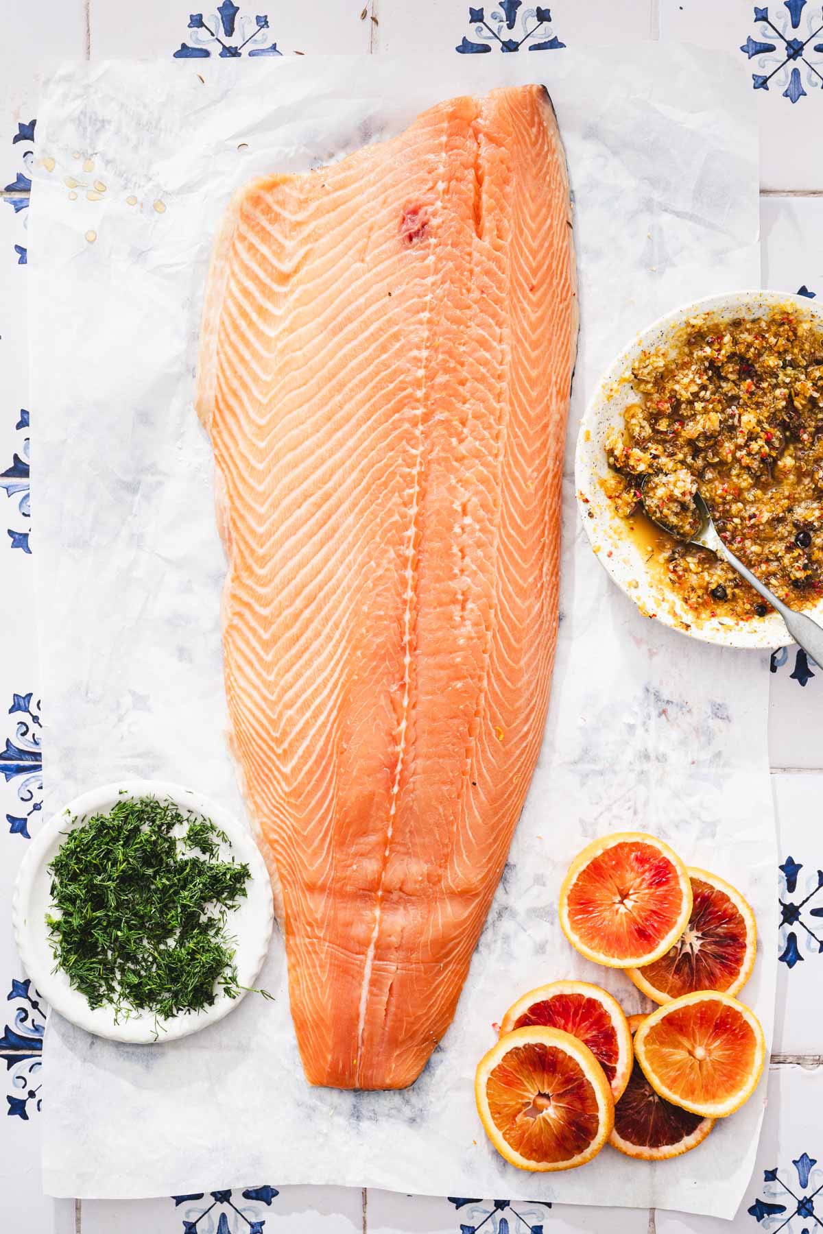 raw salmon on a parchment paper with a dish with cure, fresh dill, slices of oranges