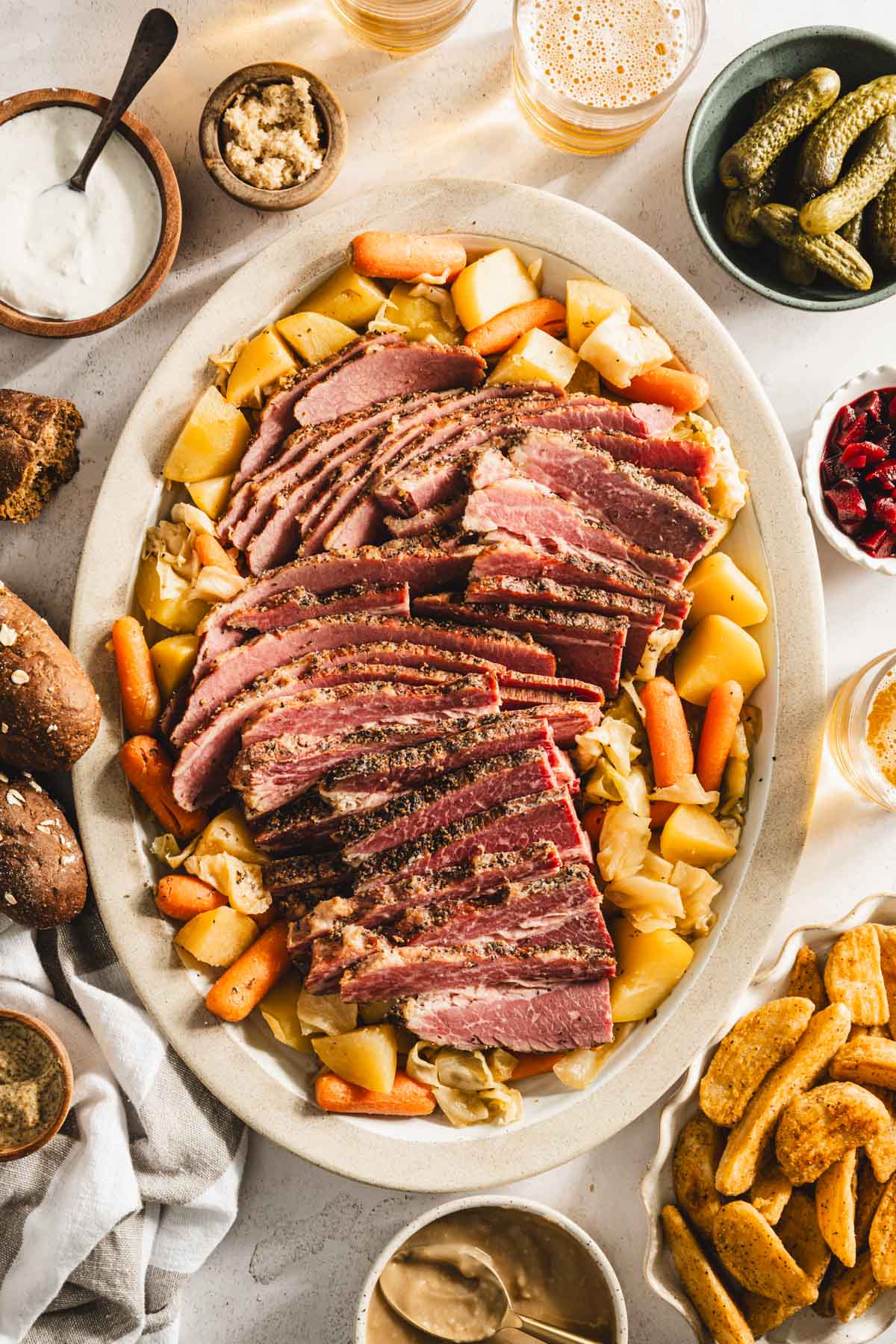 cooked corned beef brisket on a serving platter with cooked veggies underneath and to the side, glasses with beer, bowls with pickled veggies, thick cut fries, bread rolls