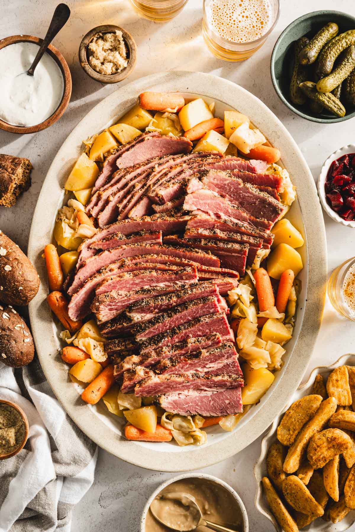 sliced cooked corned beef brisket on a serving platter with cooked veggies underneath and to the side, glasses with beer, bowls with pickled veggies, thick cut fries, bread rolls